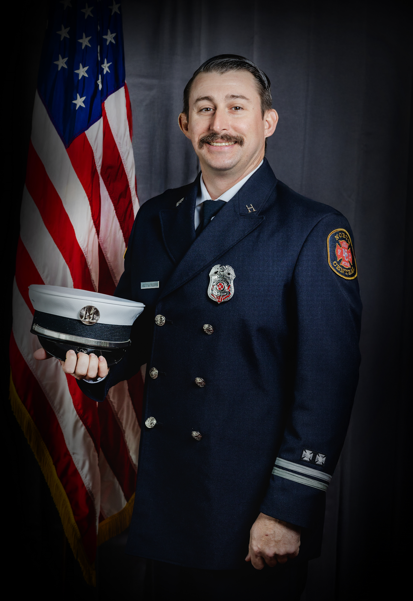 A person in a blue uniform stands smiling in front of an American flag.