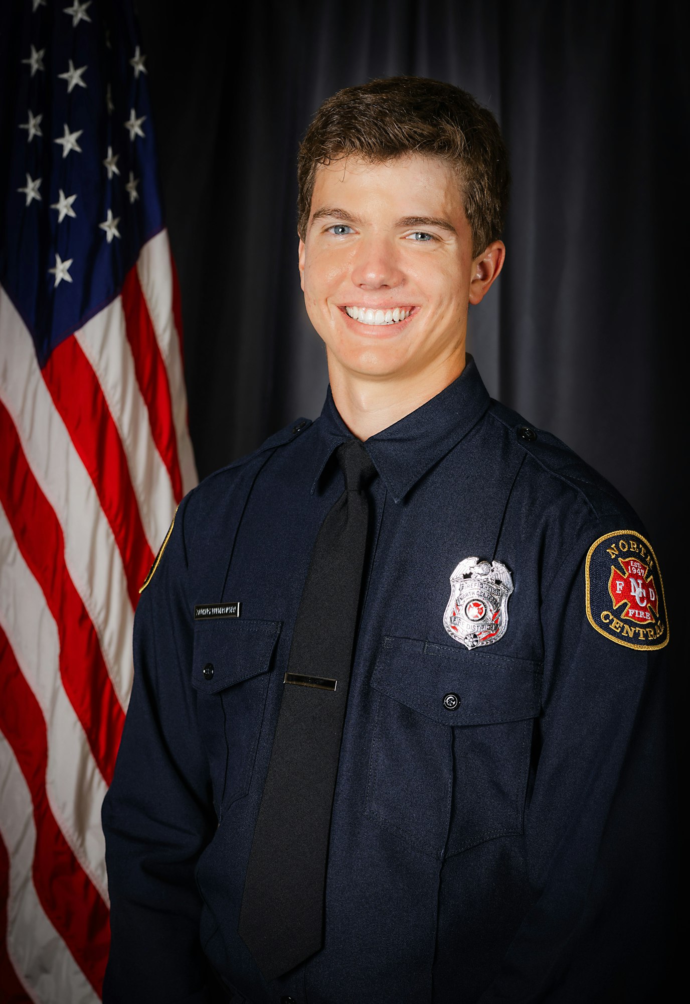 A smiling person in a firefighter's uniform with an American flag in the background.