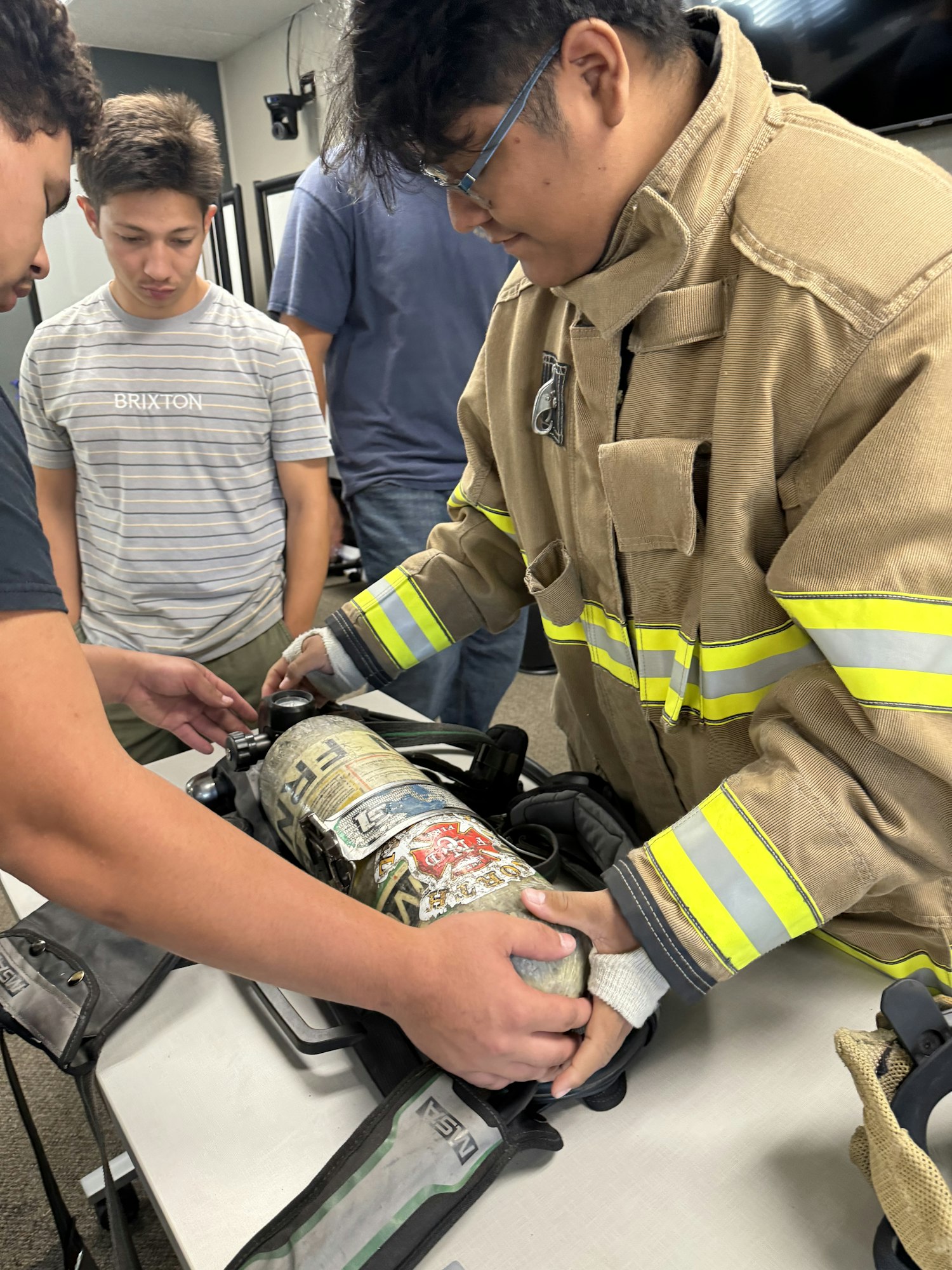People in a room examining firefighting gear, including a breathing apparatus.