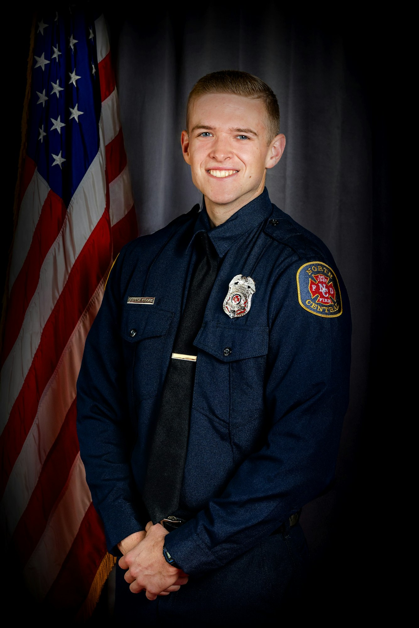 A person in a blue uniform stands smiling in front of an American flag.