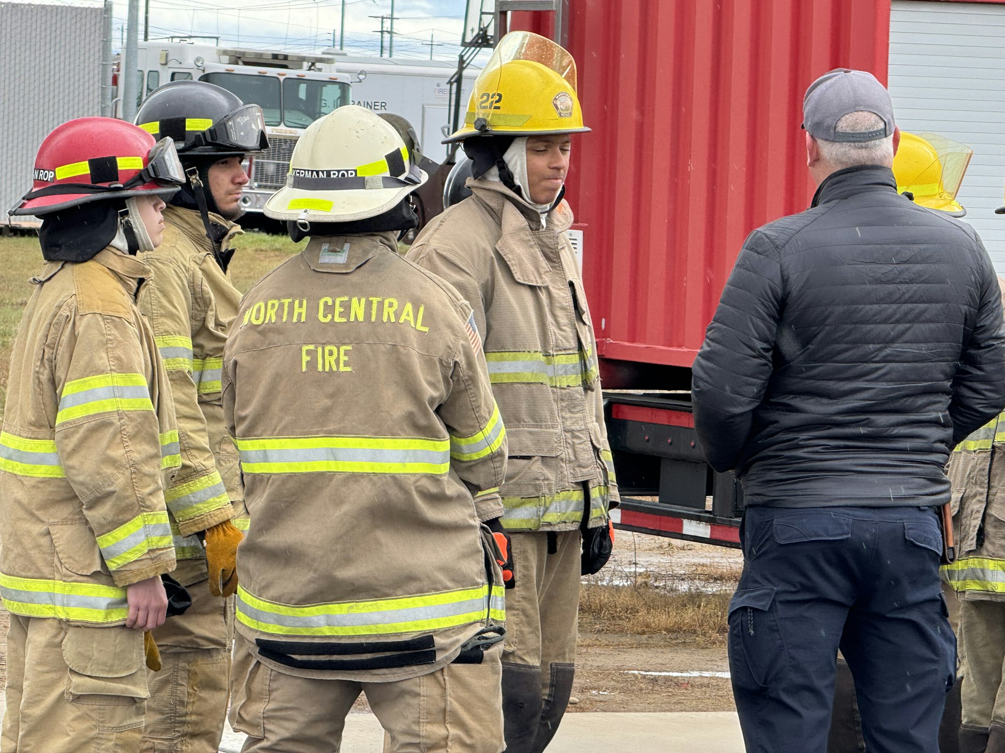 Several firefighters in gear are gathered outside near a person in a jacket, having a discussion.