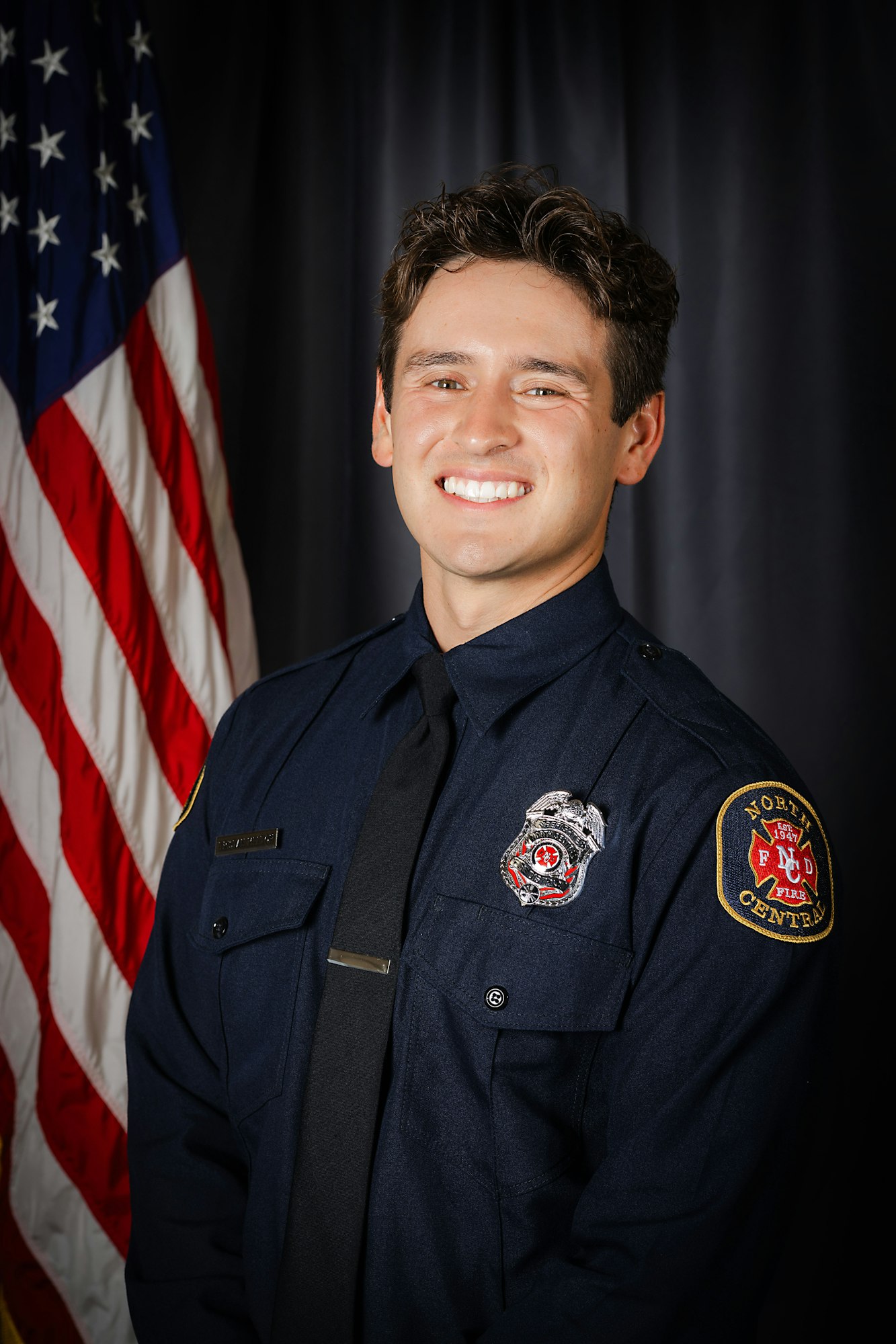 A smiling person in a firefighter uniform with an American flag in the background.