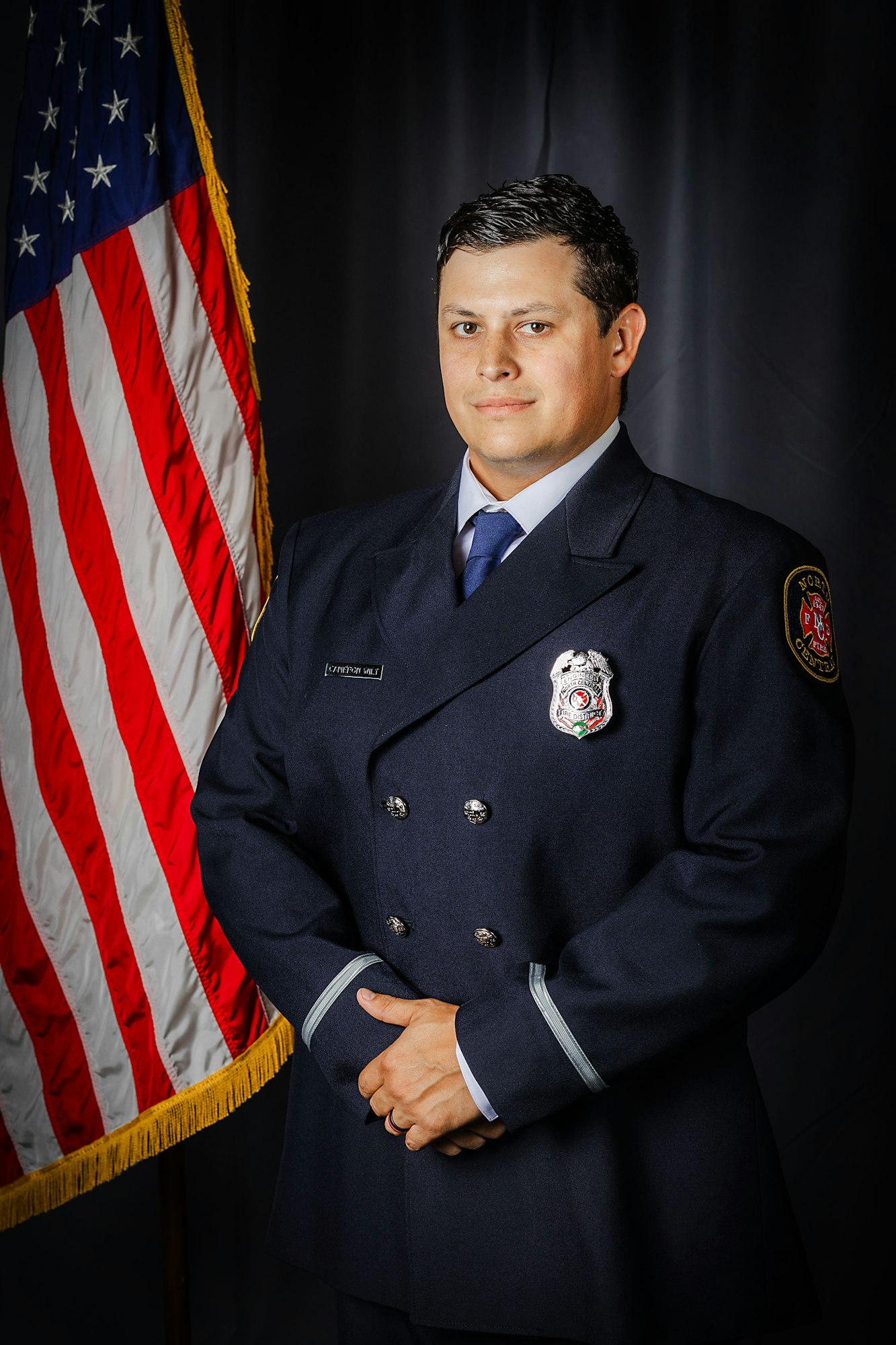 A person in a uniform stands with hands clasped in front of an American flag.