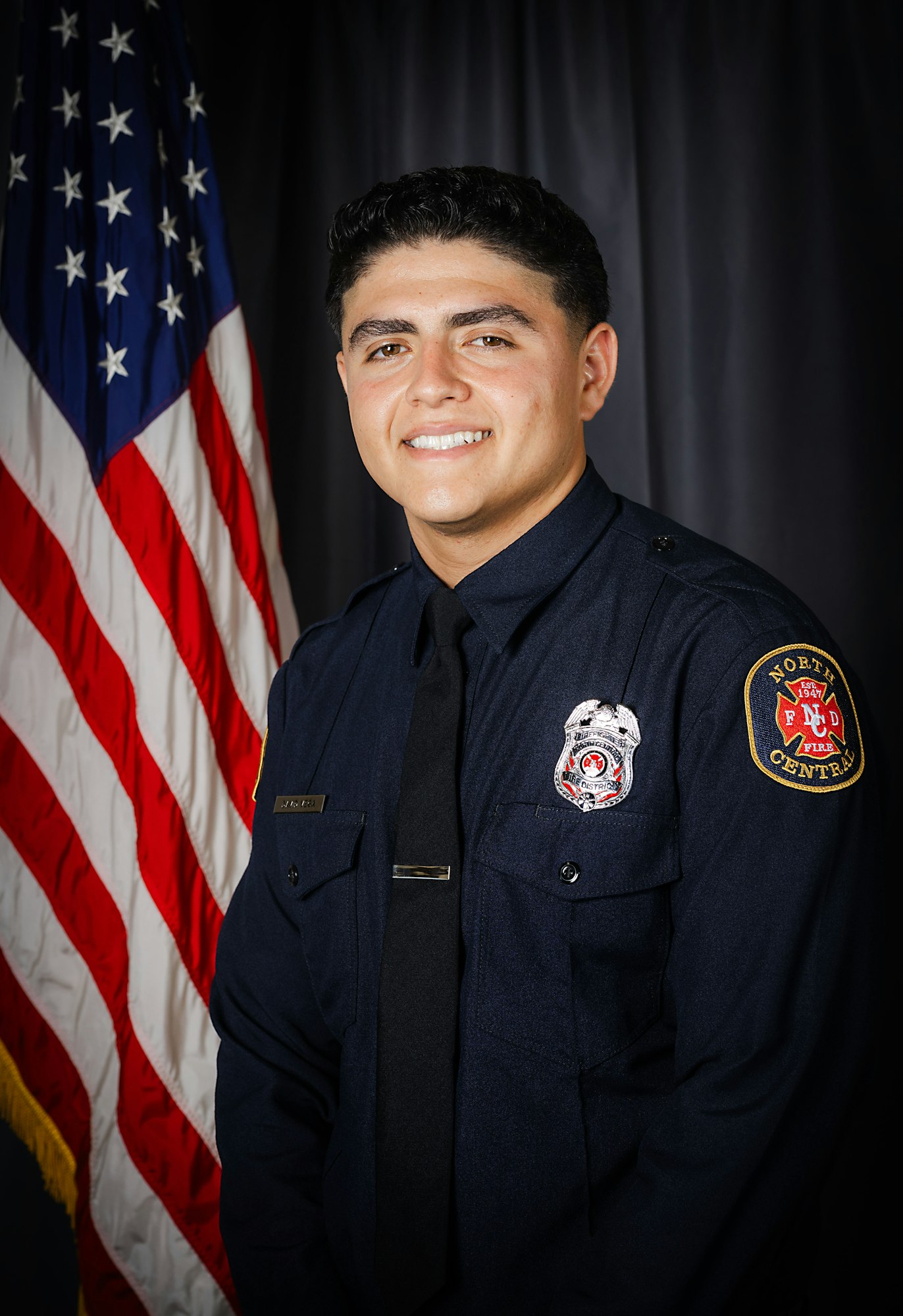 A smiling individual in a firefighter uniform with a badge, standing before an American flag.