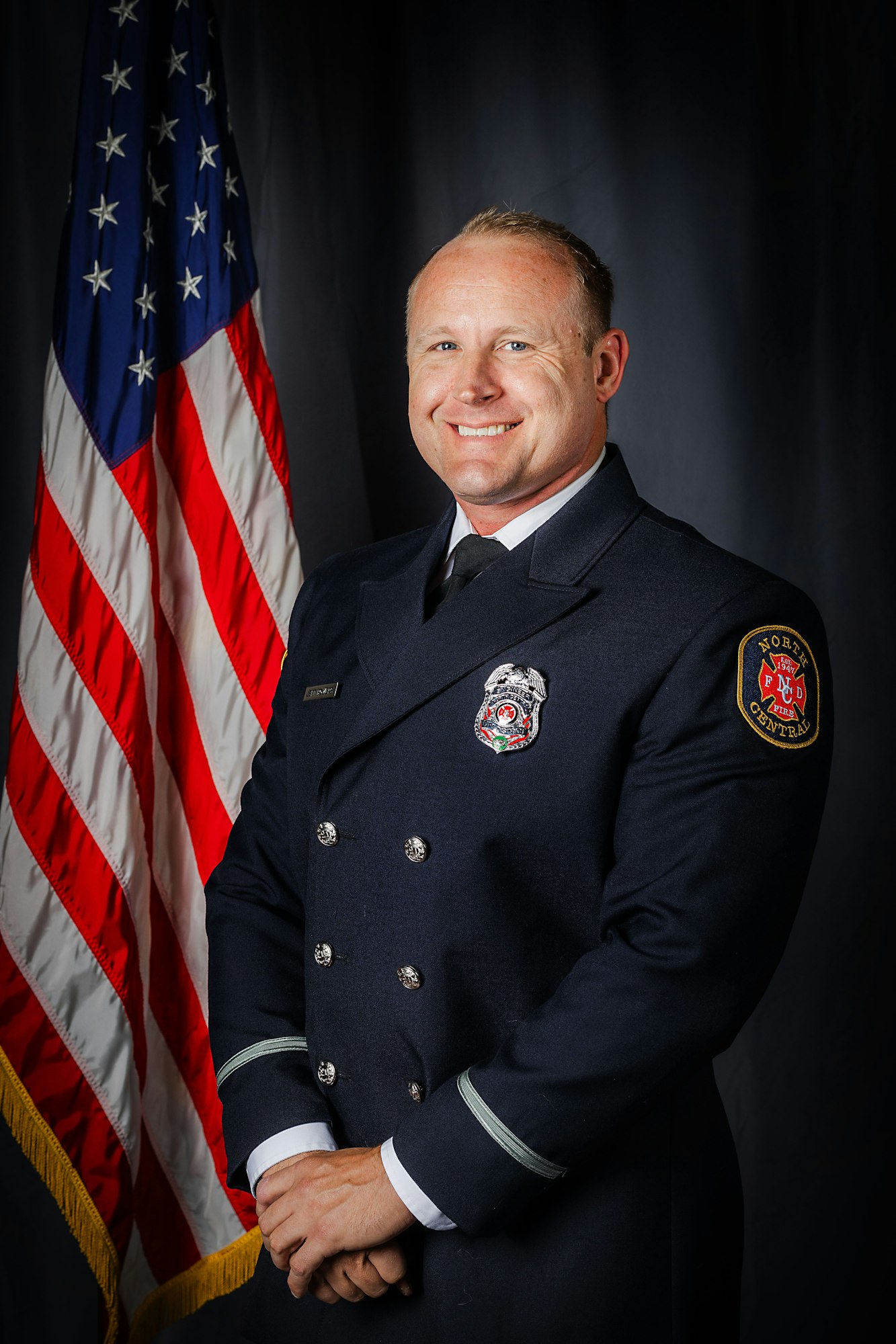 A person in a firefighter's uniform stands smiling beside a U.S. flag.