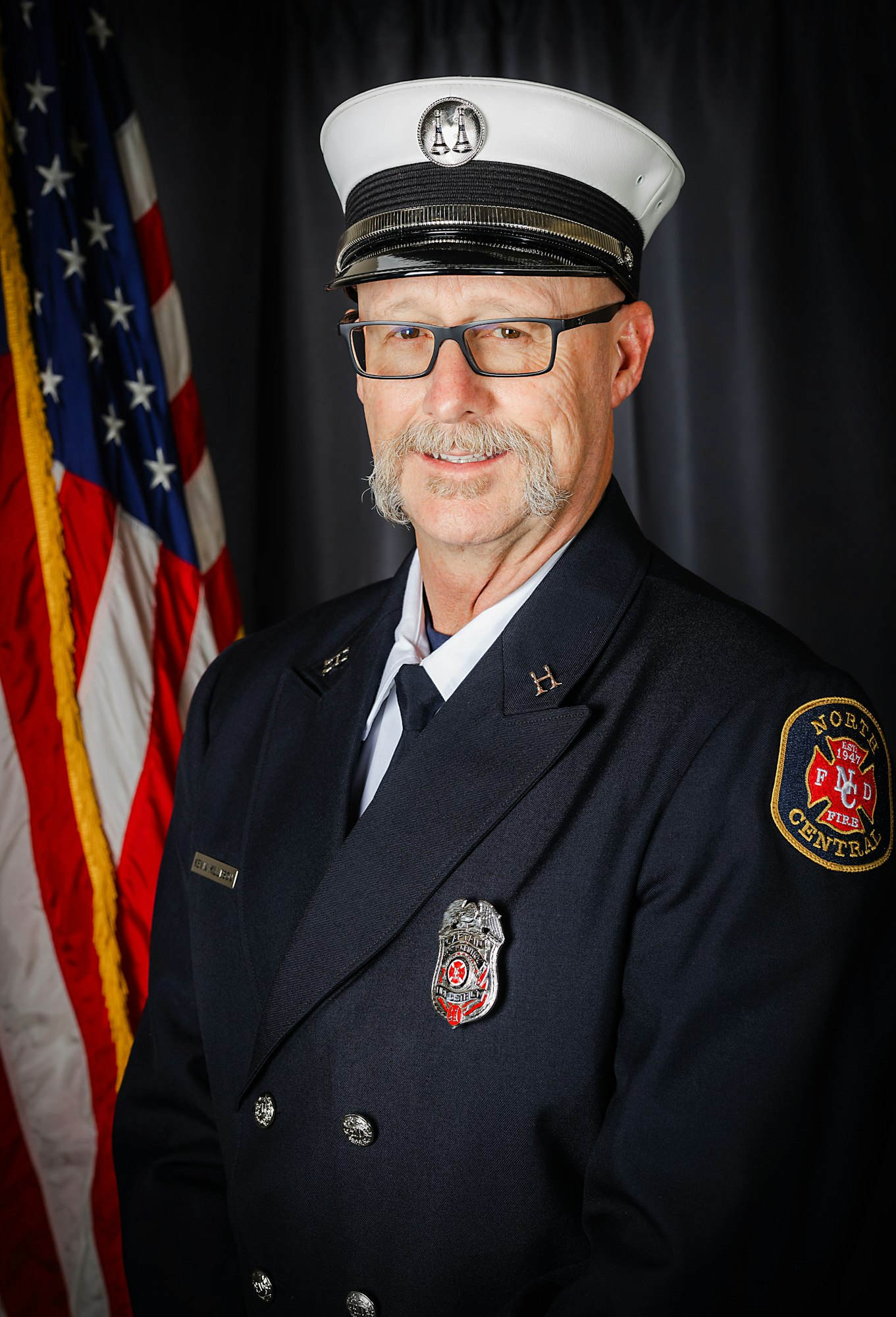 A firefighter in uniform with a white hat and glasses poses in front of a U.S. flag.