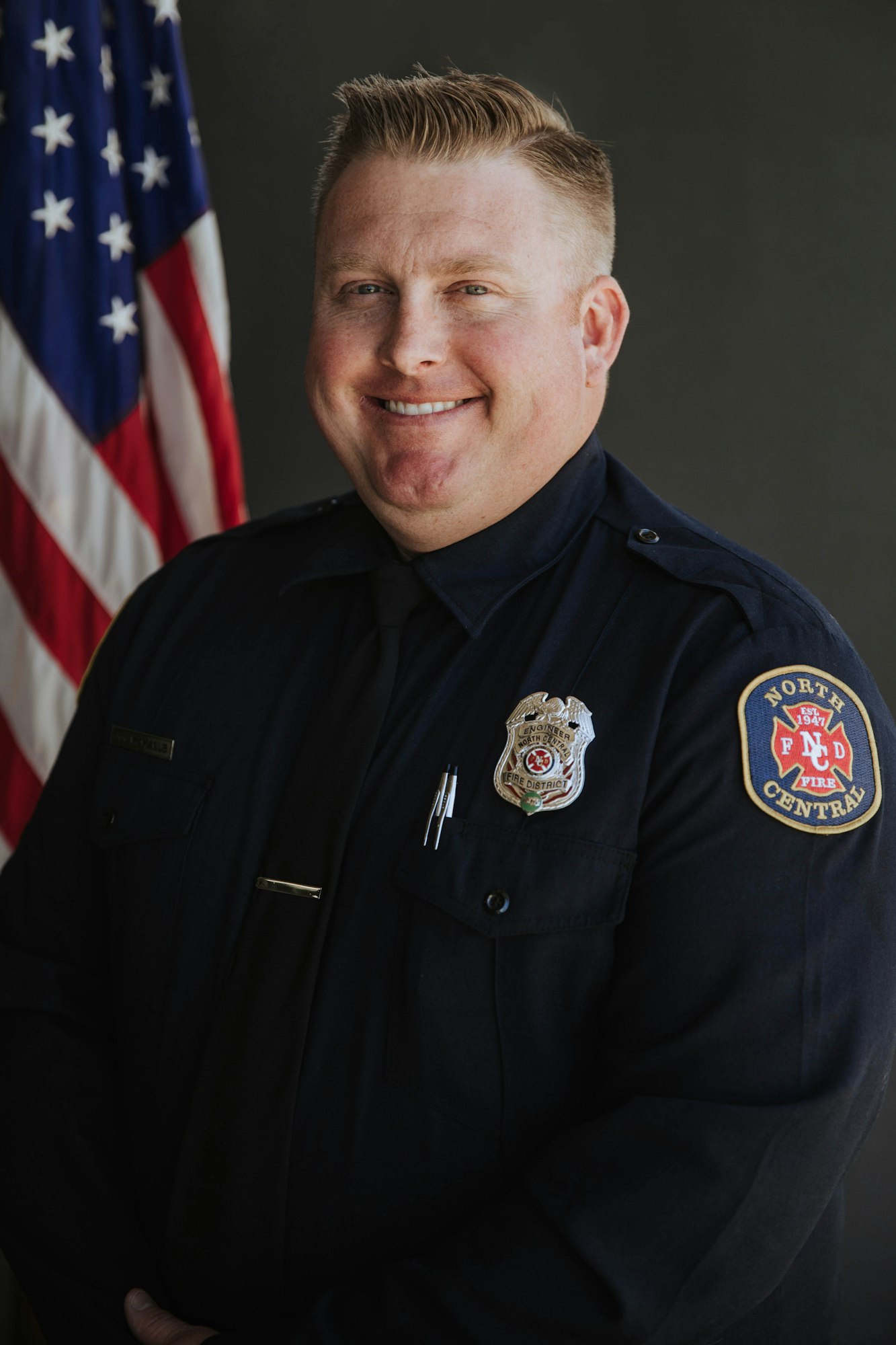 Person in a uniform with a badge, standing in front of an American flag.