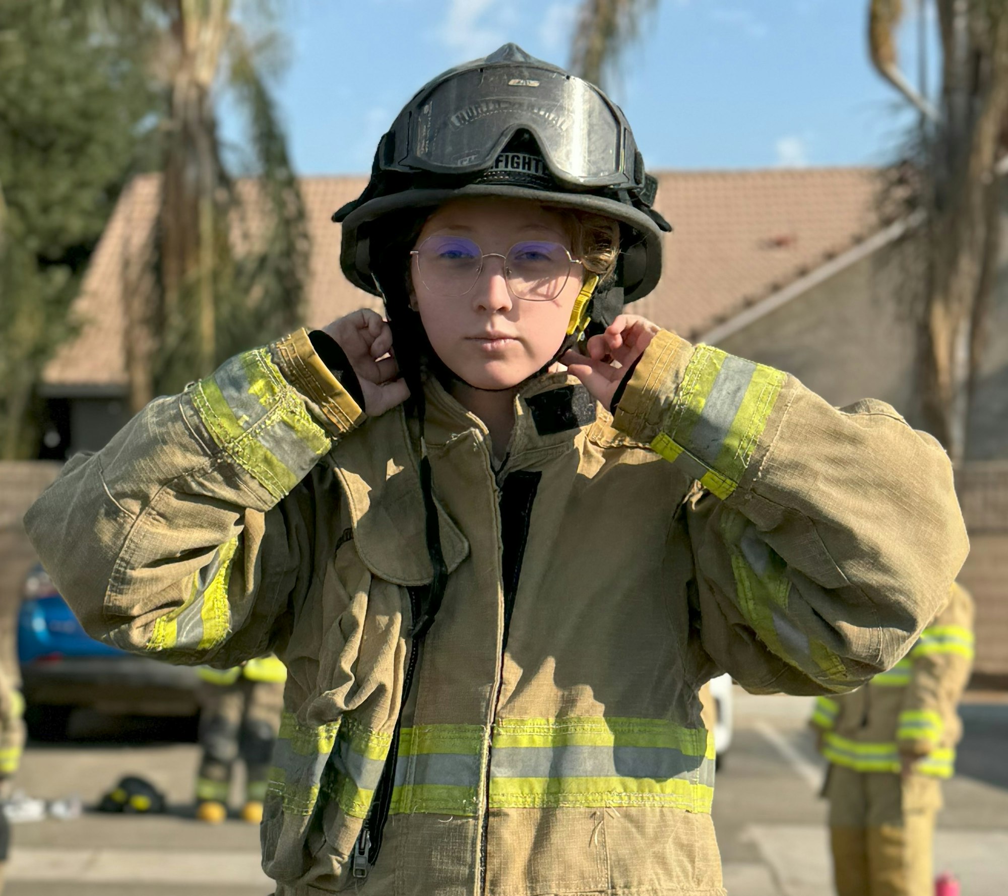 Person wearing firefighter gear, adjusting their helmet, with a house and trees in the background.