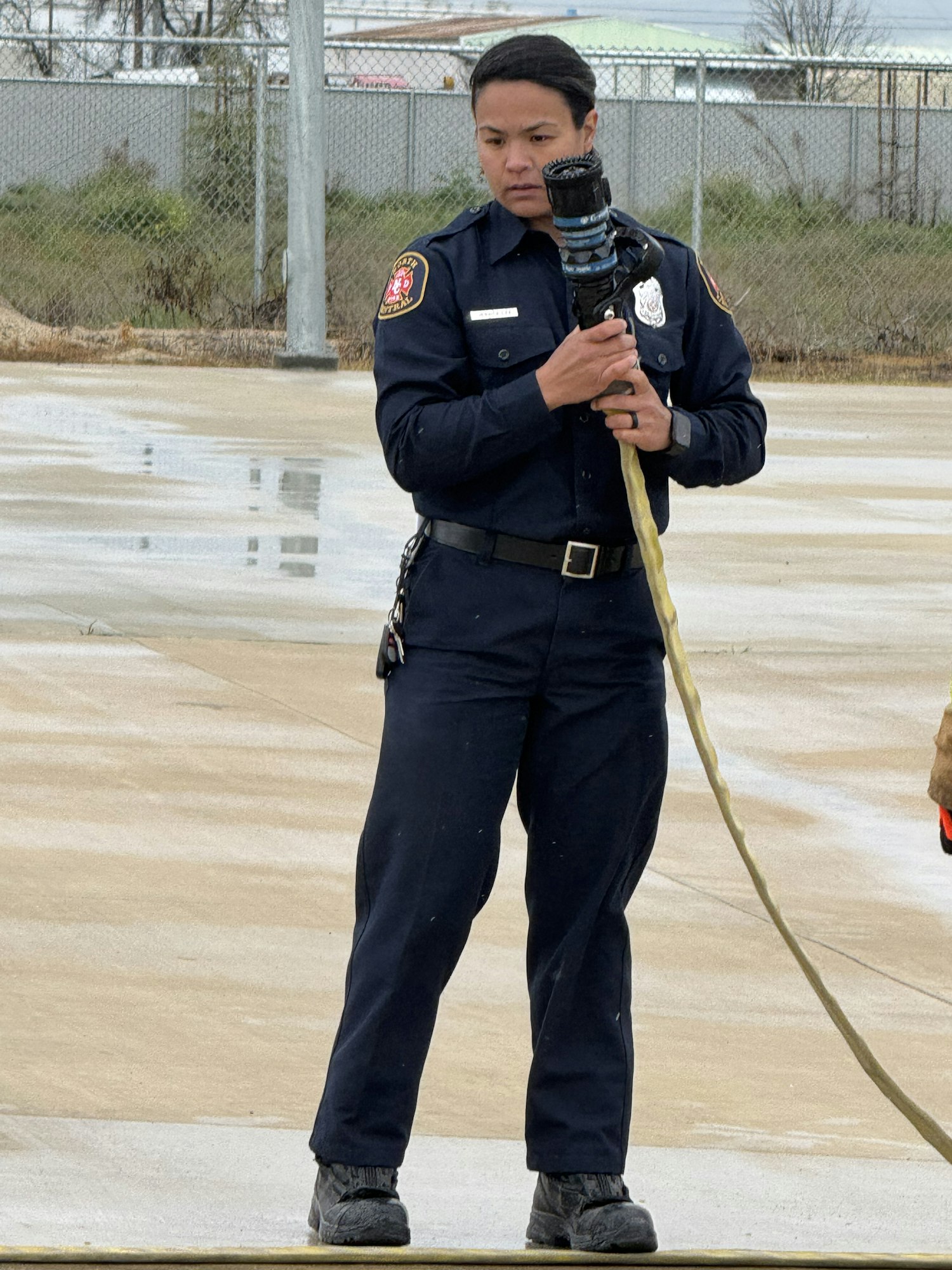 A person in a firefighter uniform holds a hose, standing on a wet pavement.
