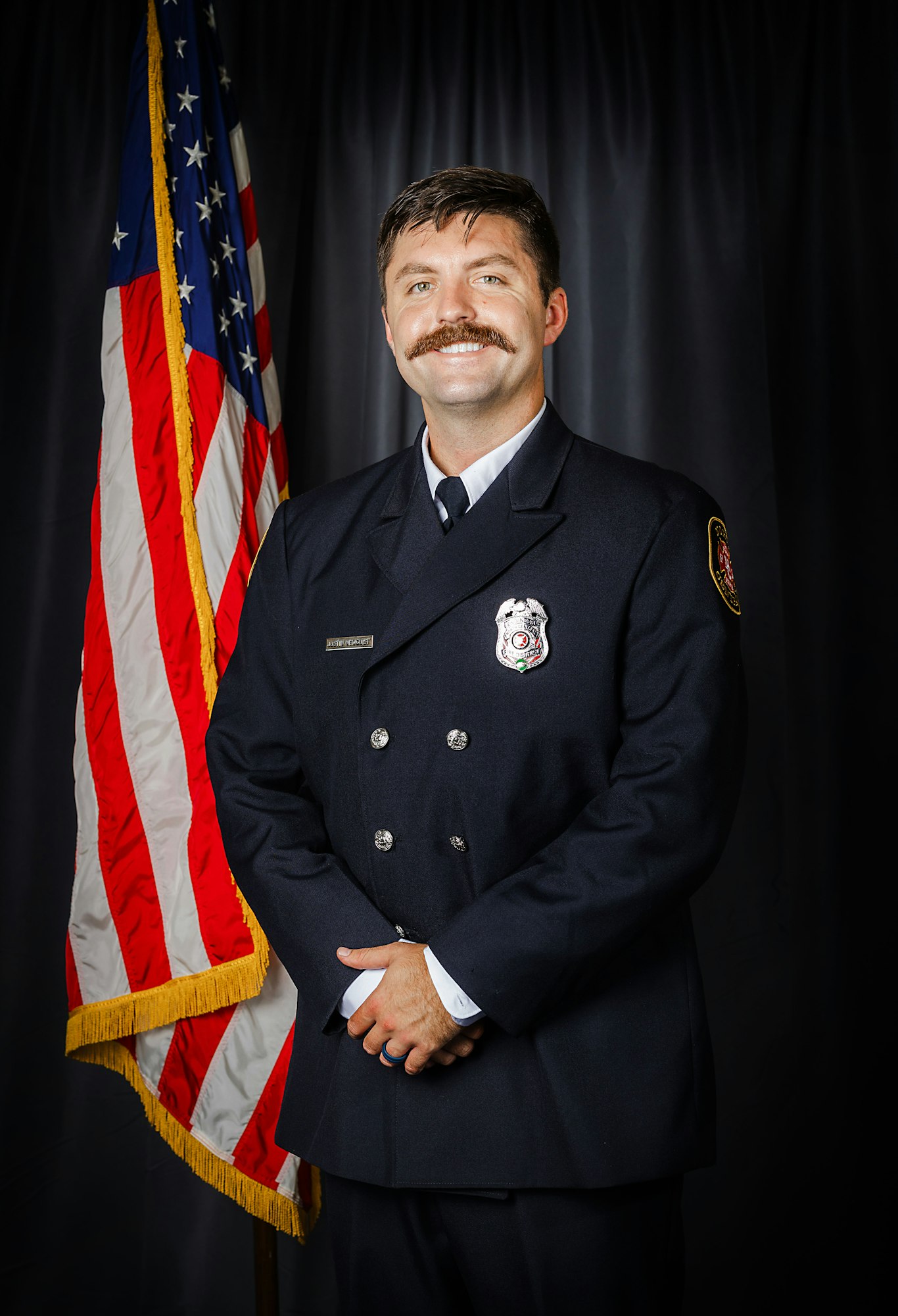 Fire Engineer Newquist in uniform smiling, standing before an American flag.
