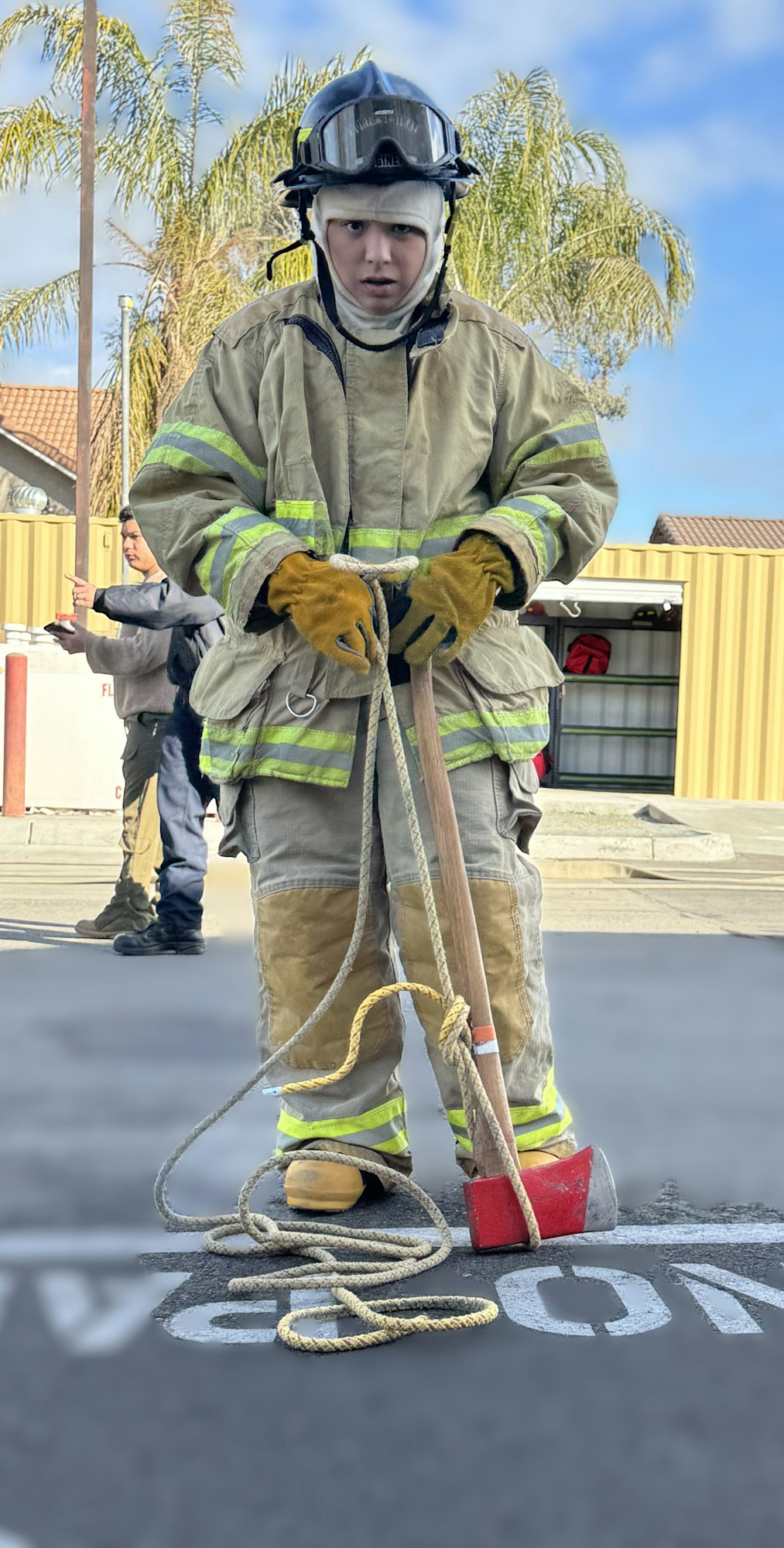 Person in firefighter gear holding an axe and rope, outdoors.