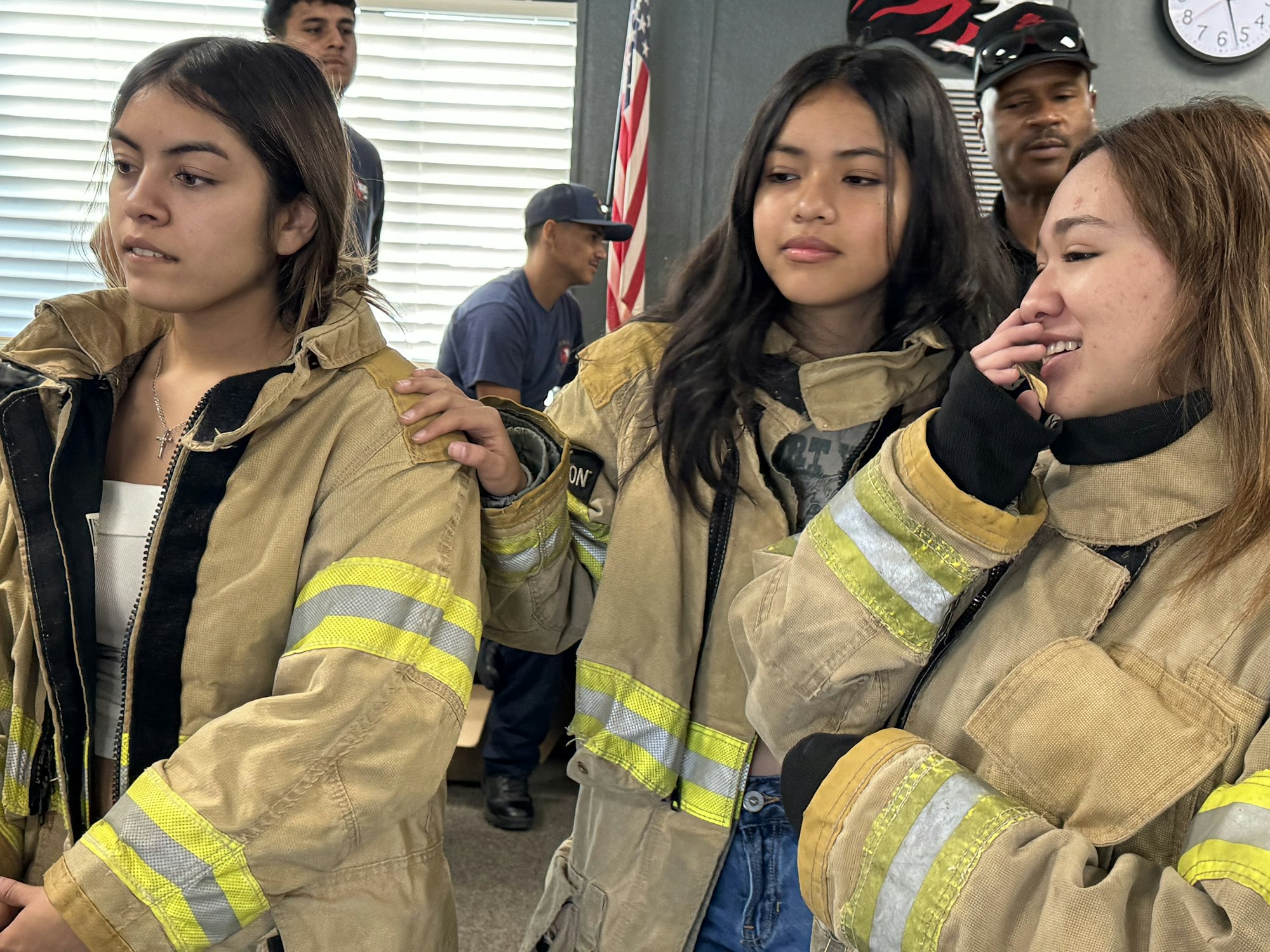 Three people wearing firefighter jackets, standing indoors, with two people in the background.