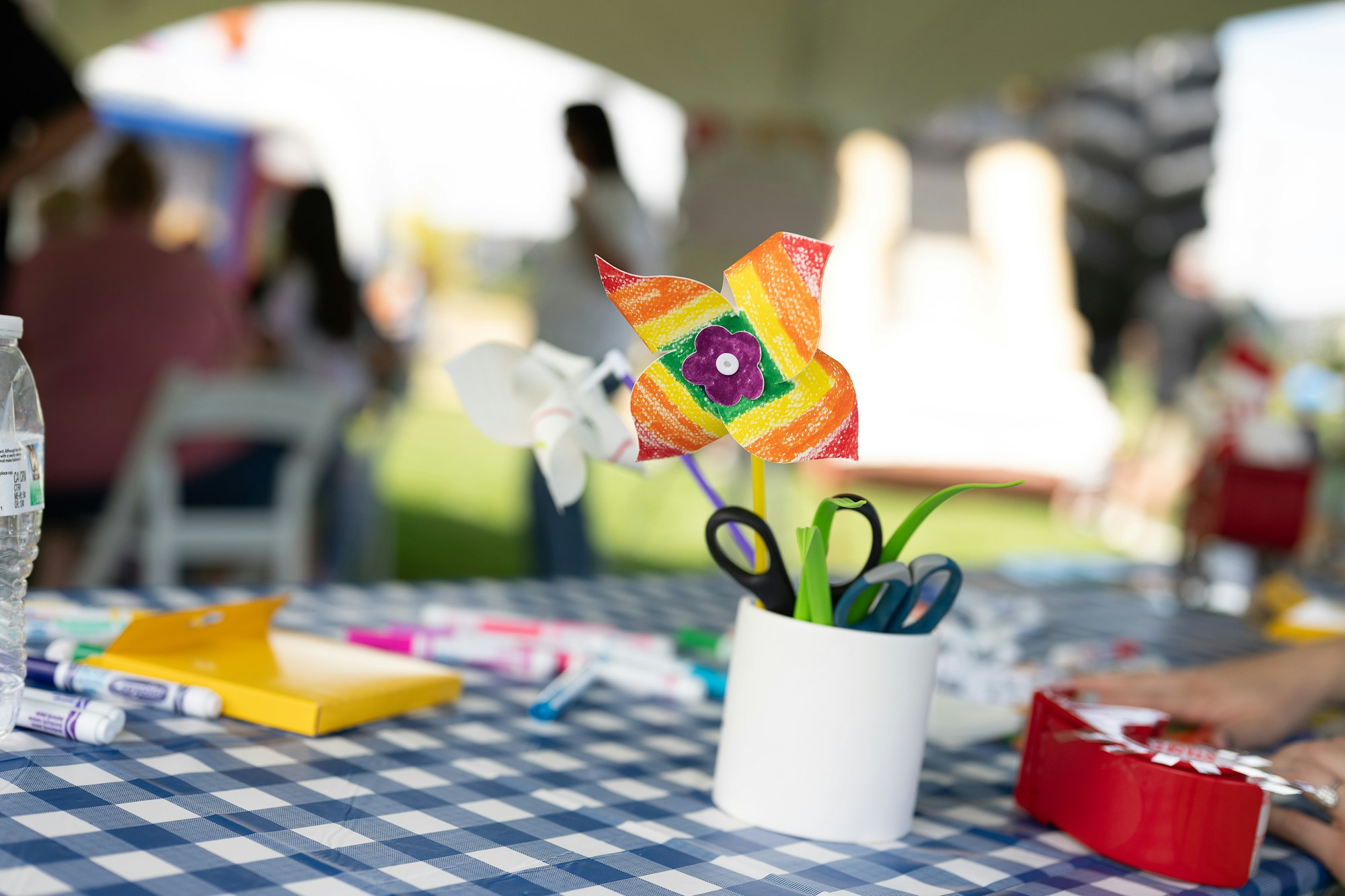 A colorful paper pinwheel in a cup with scissors on a checkered tablecloth, markers, and a water bottle.
