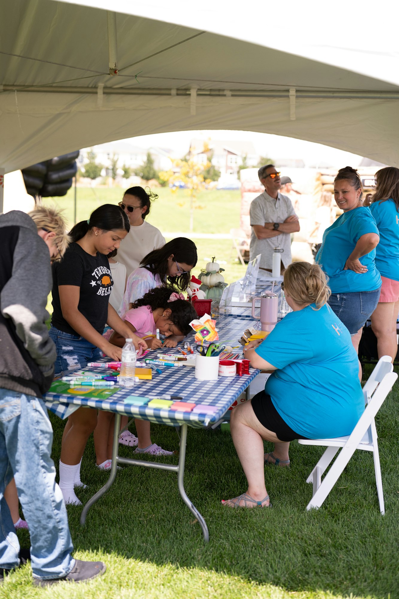 People are gathered under a tent, engaged in arts and crafts at a table covered with a checkered cloth.