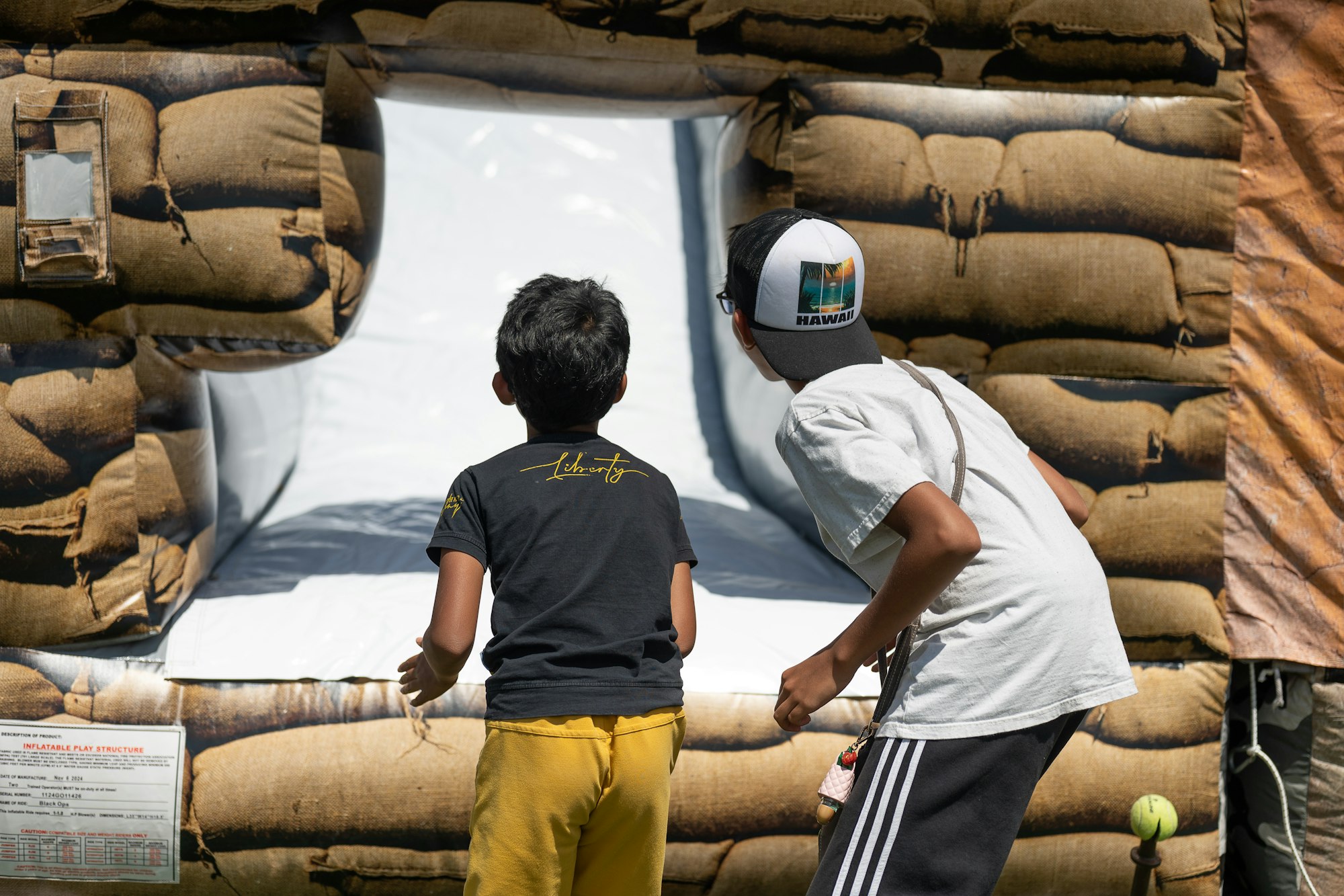 Two kids looking at an entrance of an inflatable structure with a white slide, surrounded by brown sack-like patterns.