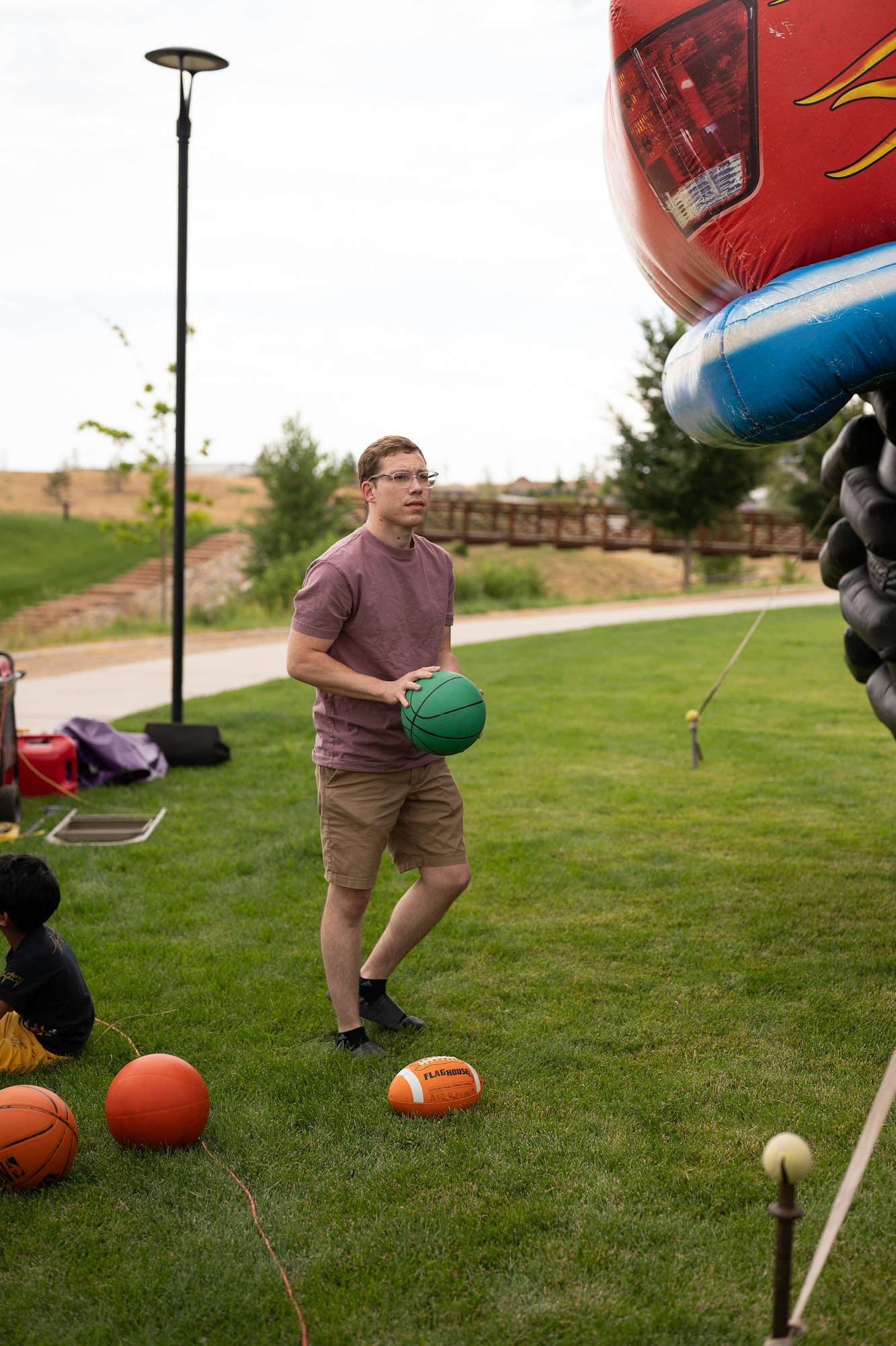 A person holds a green ball near an inflatable structure, with basketballs and a football on the grass at an outdoor park.