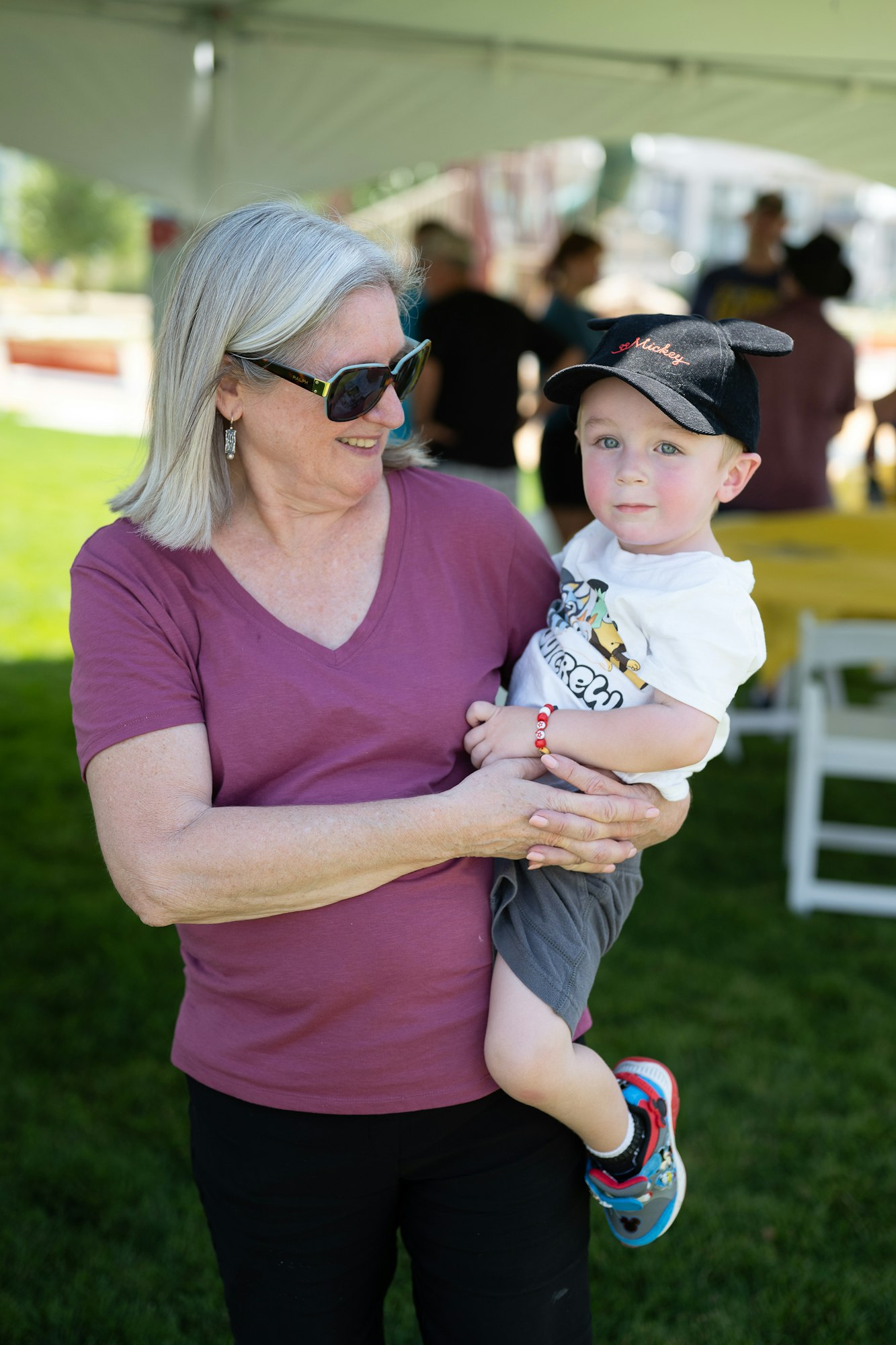 A woman wearing sunglasses holds a child wearing a Mickey Mouse cap in a sunny outdoor setting.