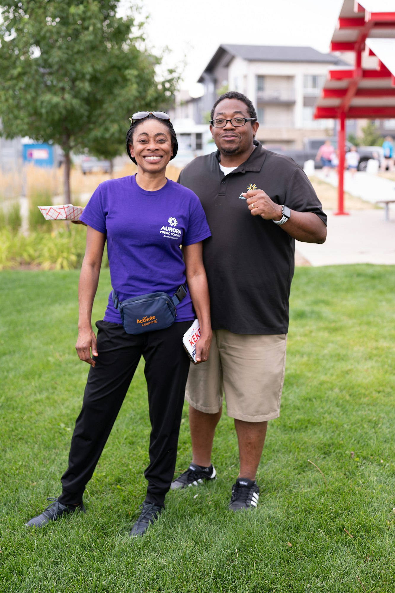 Two people smiling, standing on grass. One in a purple "Aurora Public Schools" shirt, the other in a black shirt with text.