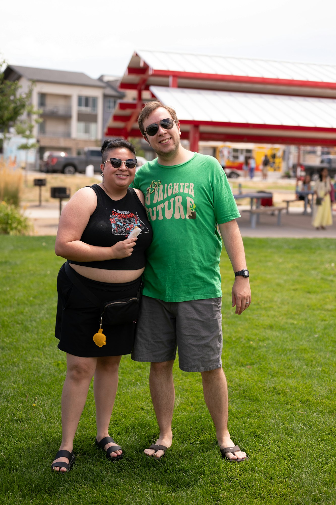 Two people smiling, wearing sunglasses, standing on grass in front of a red structure and buildings.