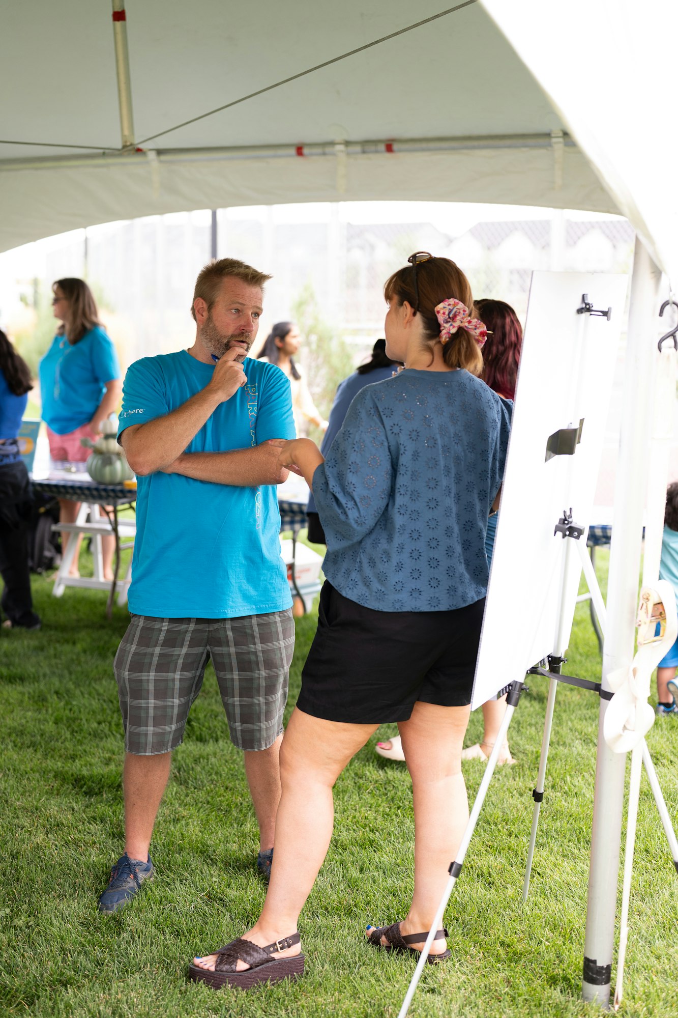 Two people talking under a tent at an outdoor event. Other attendees and a table are in the background.