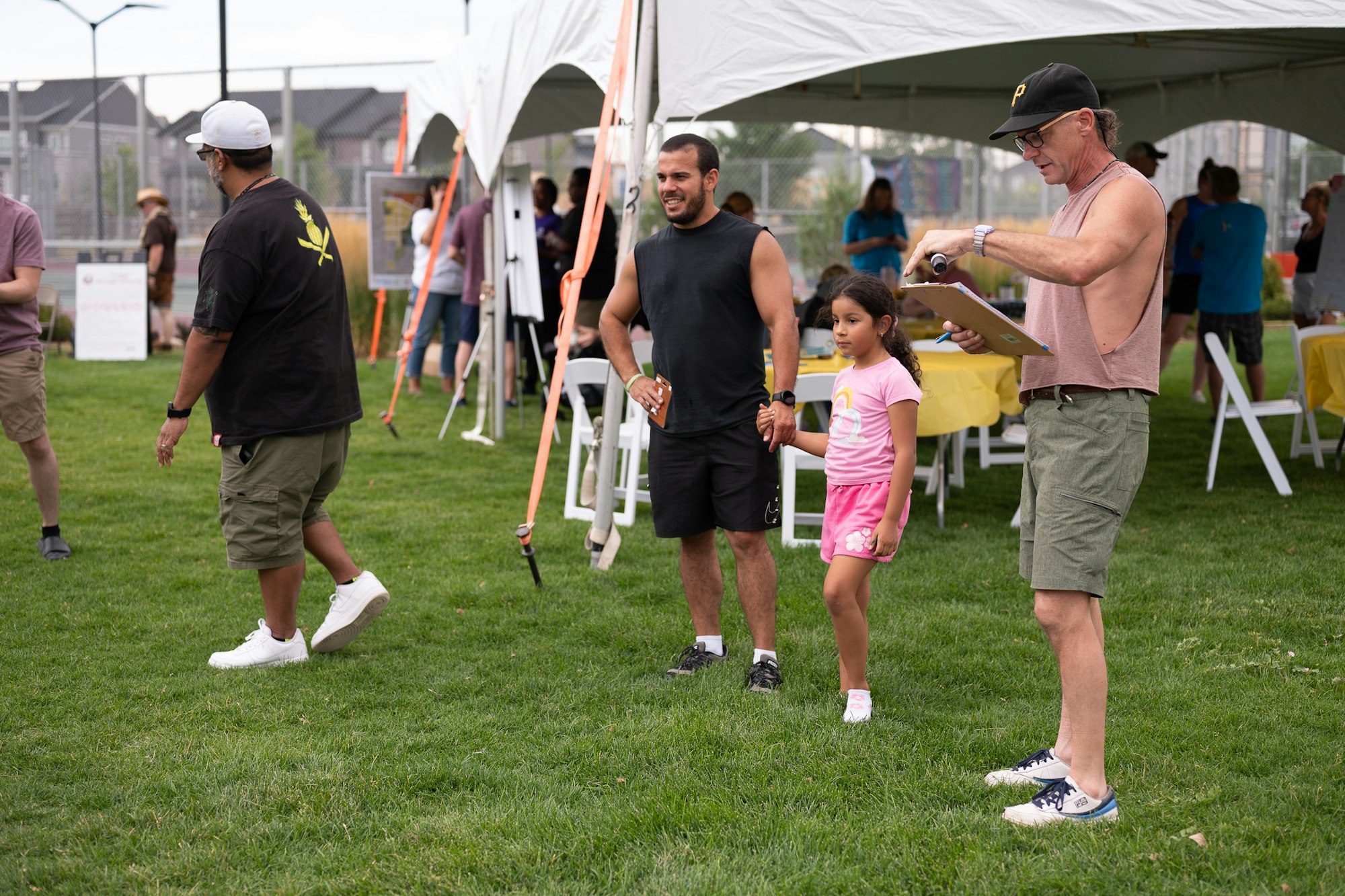 People at an outdoor event with tents, a man with a clipboard, and a child holding a man's hand on a grassy field.