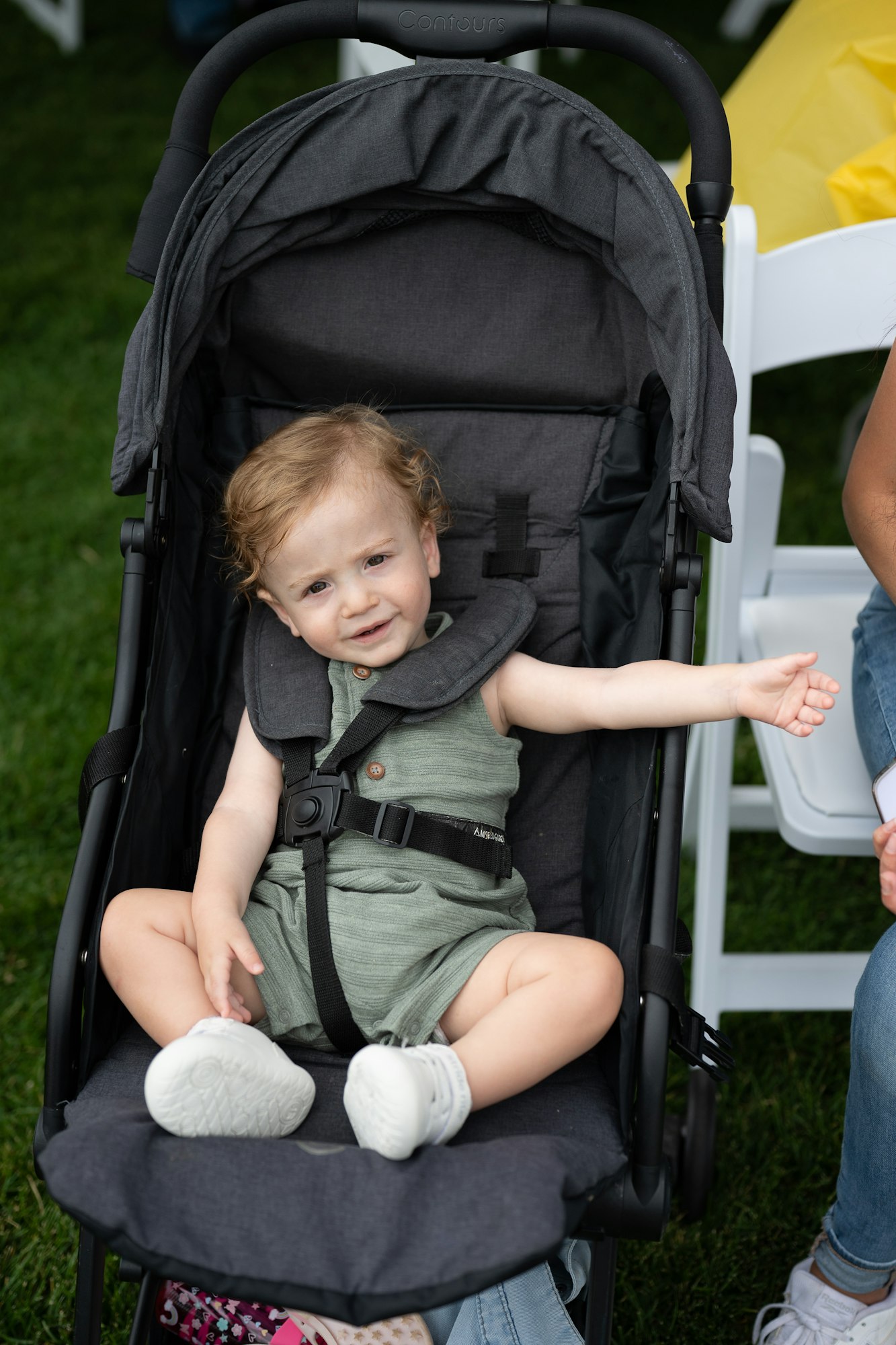 A toddler sitting in a stroller on grass, wearing a green outfit and white shoes, smiling with an outstretched arm.