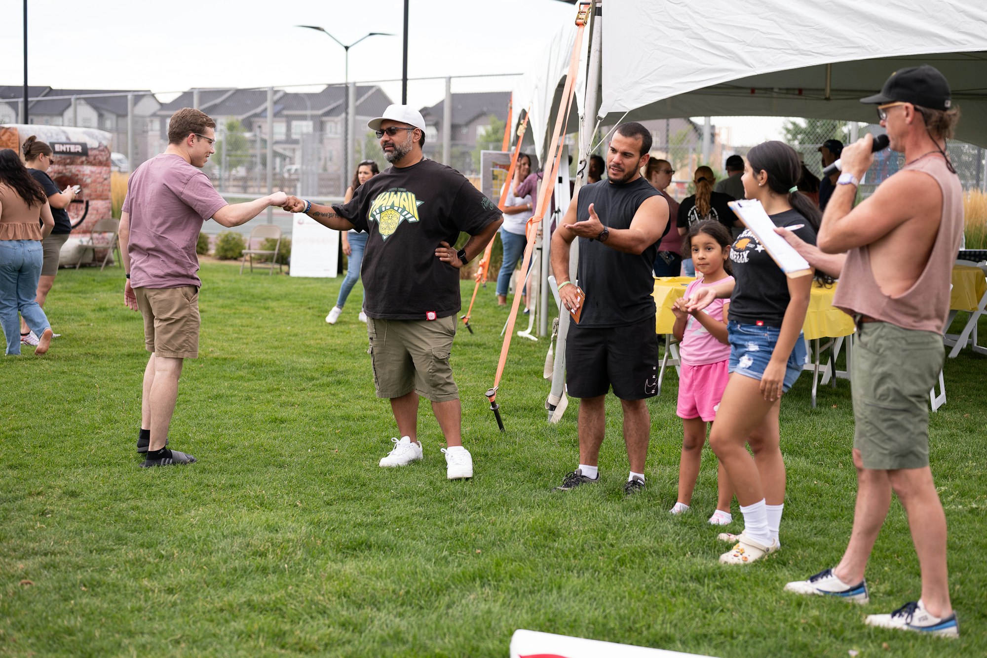People interacting at an outdoor event with tents and a speaker. One person is holding a microphone and another fist-bumps.