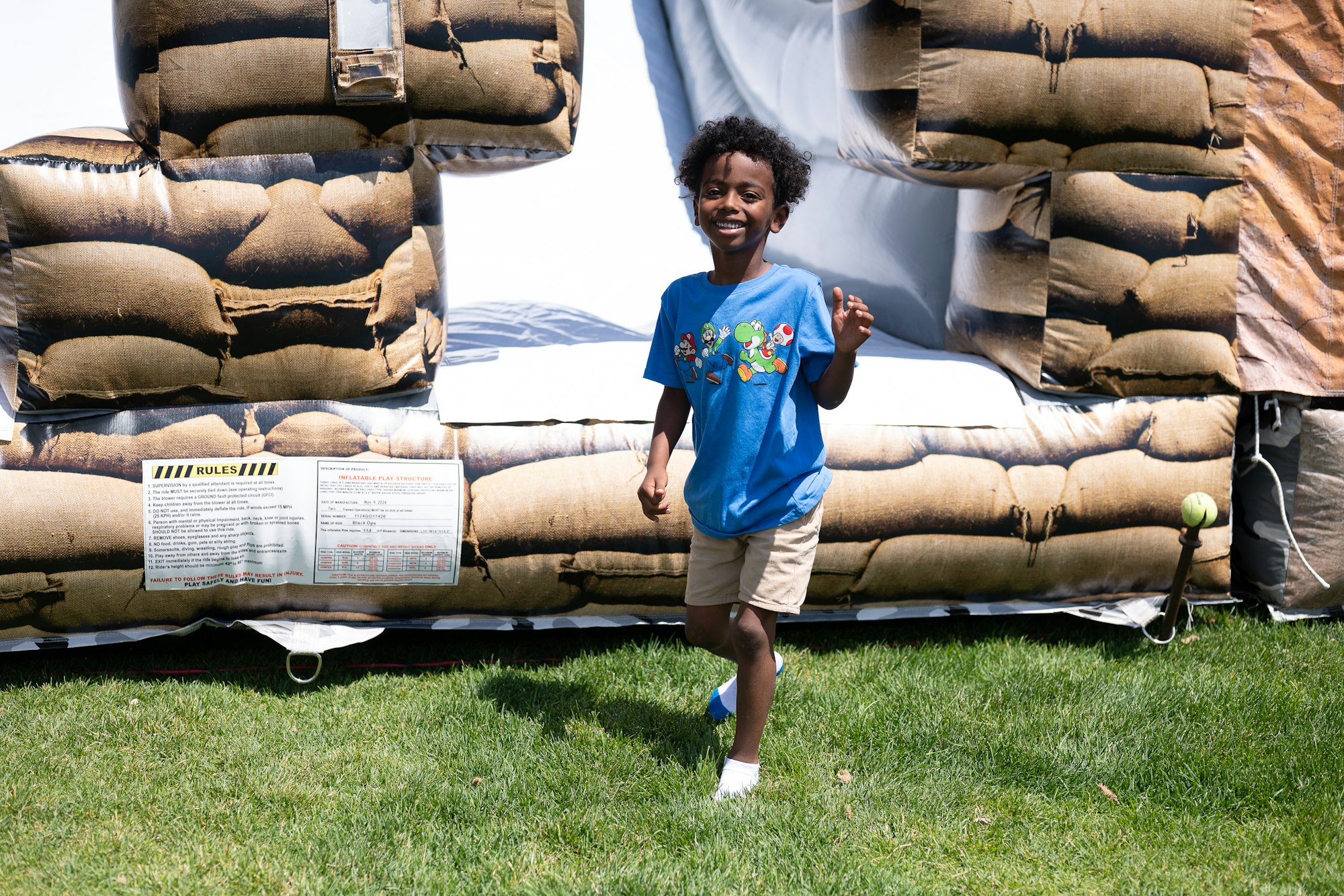 A child in a blue shirt stands in front of an inflatable with sandbag design, smiling on a grassy area.