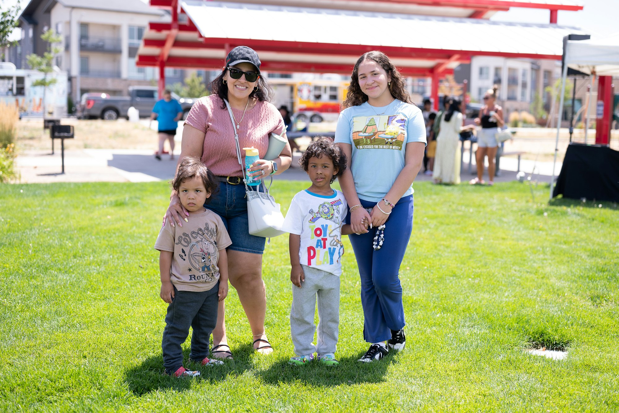 A group of four people, two adults and two children, standing on grass in a park with buildings in the background.
