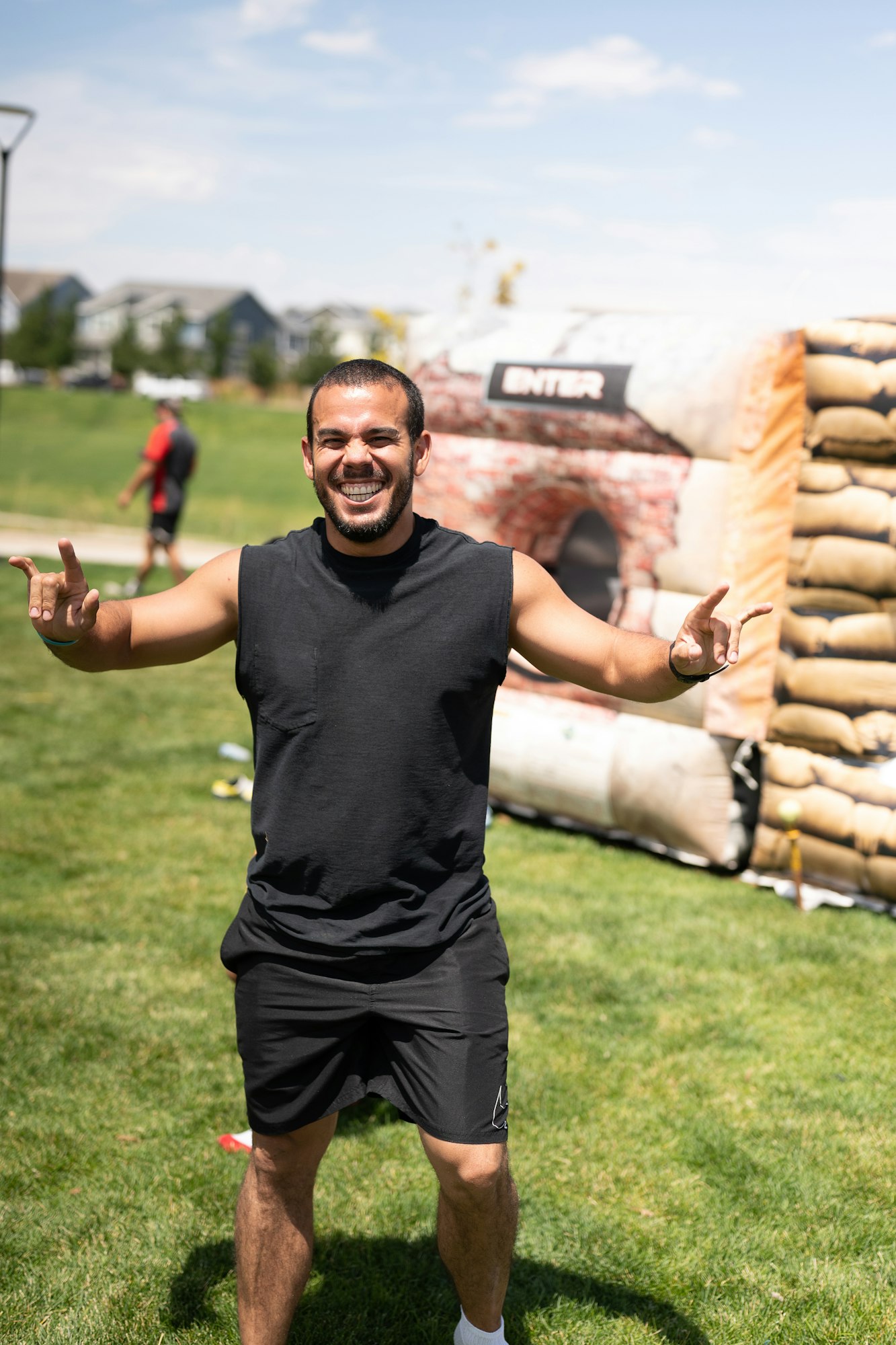 A smiling person outdoors, wearing a black sleeveless shirt and shorts, making a gesture with both hands. An inflatable structure labeled "ENTER" is in the background.