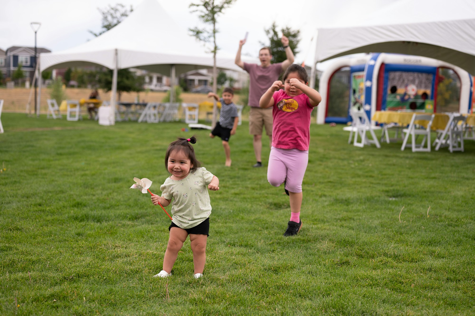 Children playing on grass with a pinwheel, under white tents; adults and another child in the background.