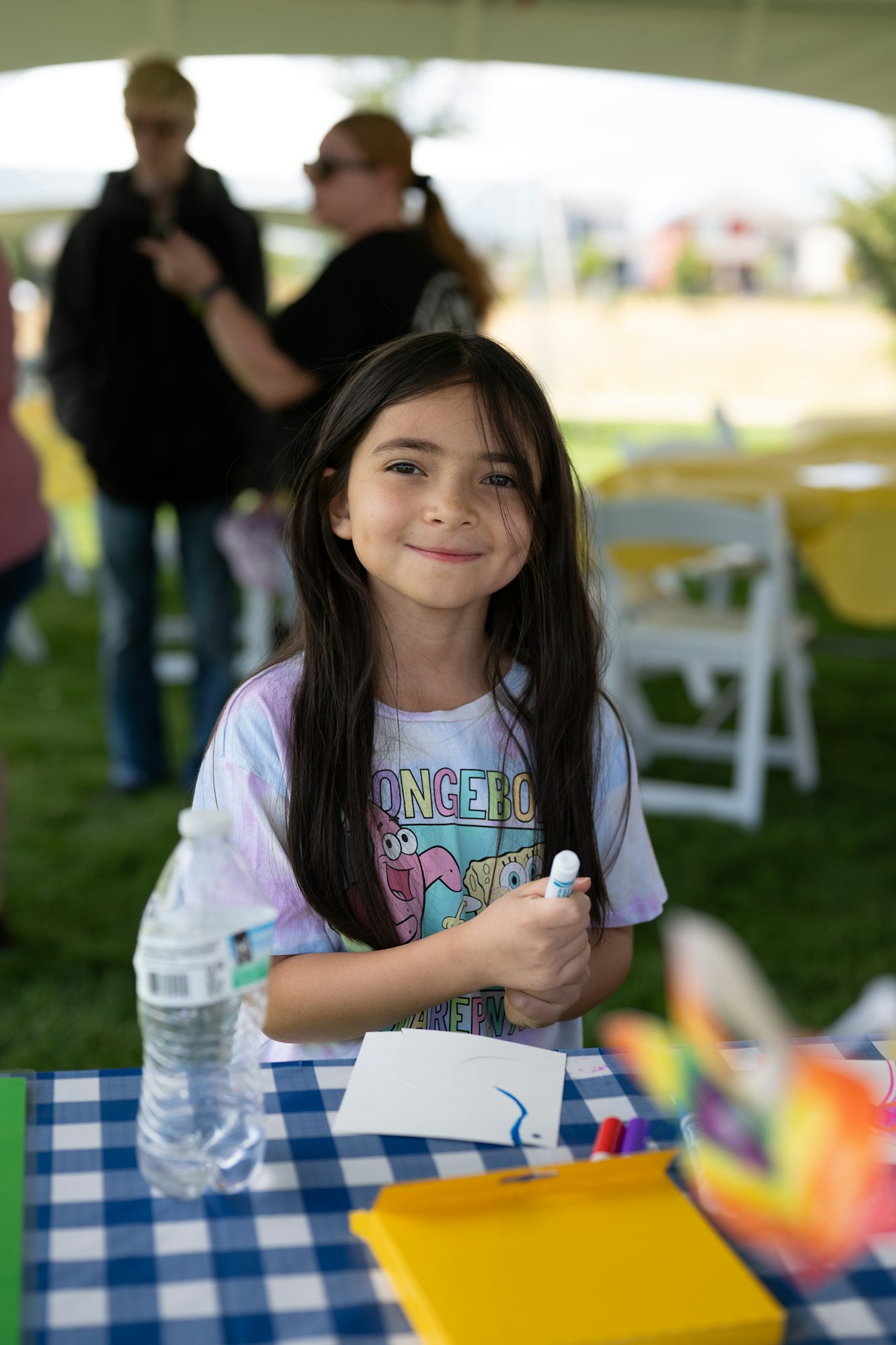 A girl at an outdoor event, drawing with a marker at a table, wearing a colorful shirt.