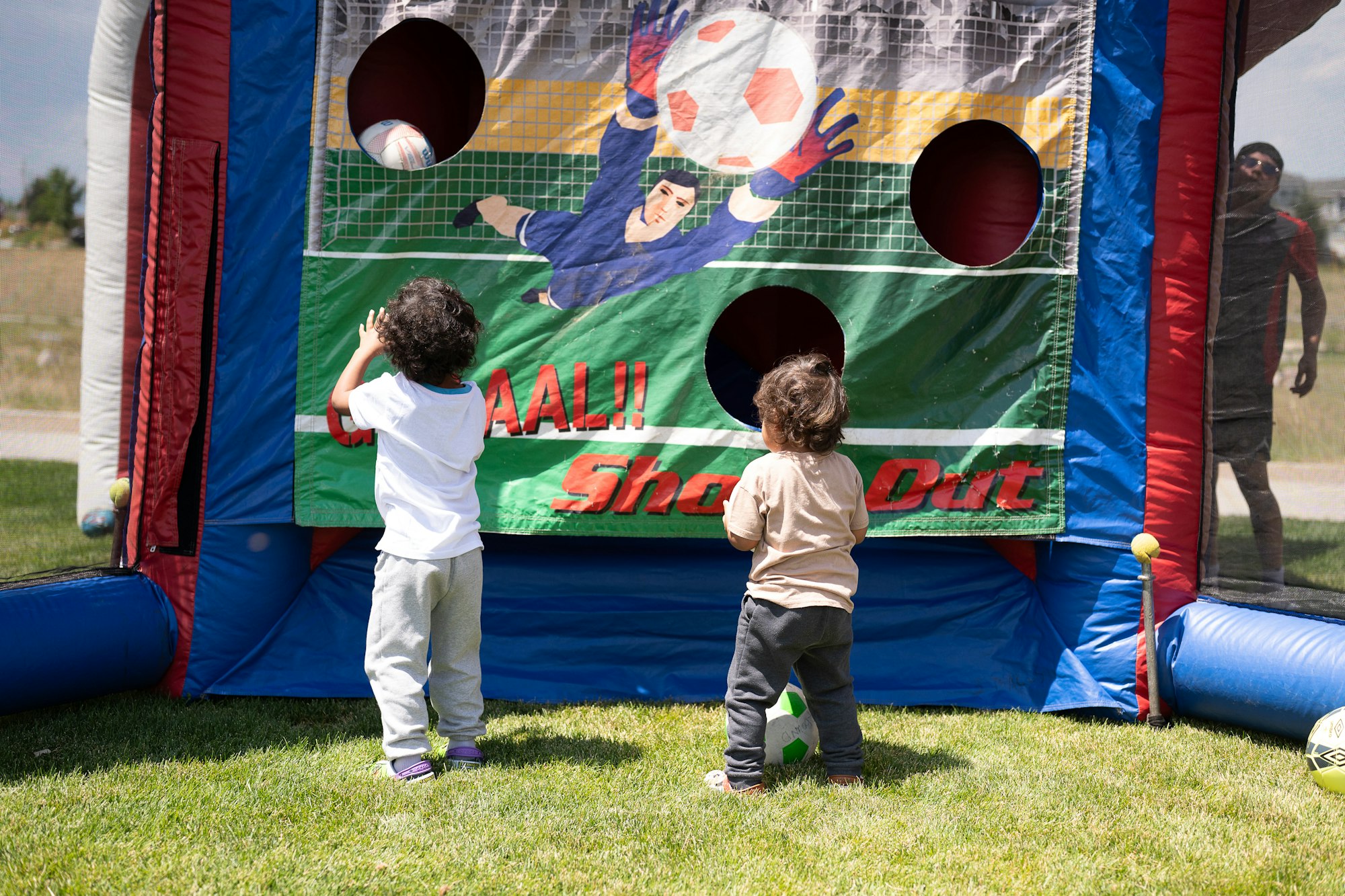 Two children playing with a soccer game inflatable featuring a goalie theme with balls on the grass.