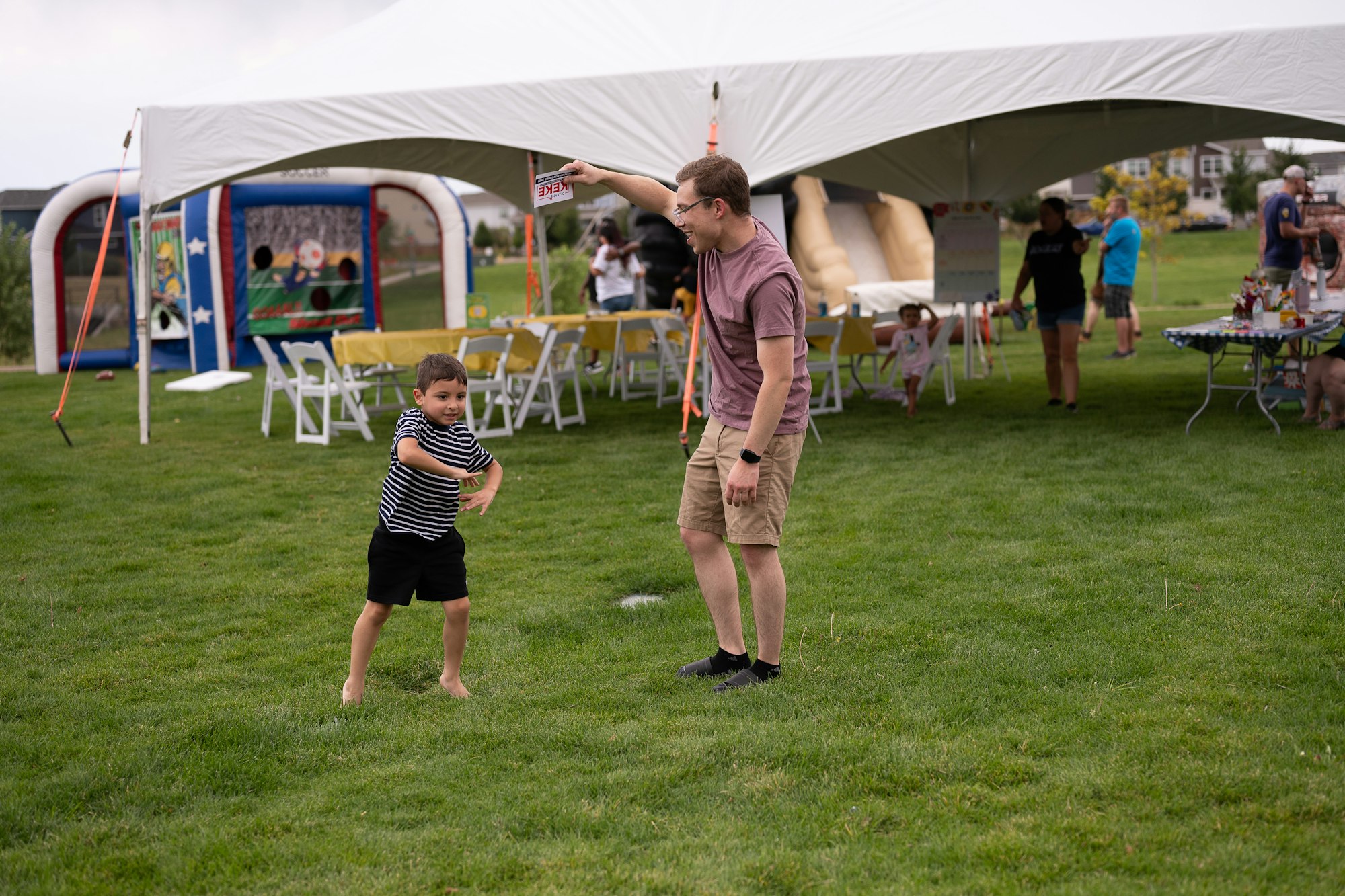 A child dances on grass beside an adult under a tent, with party tables and an inflatable game in the background.