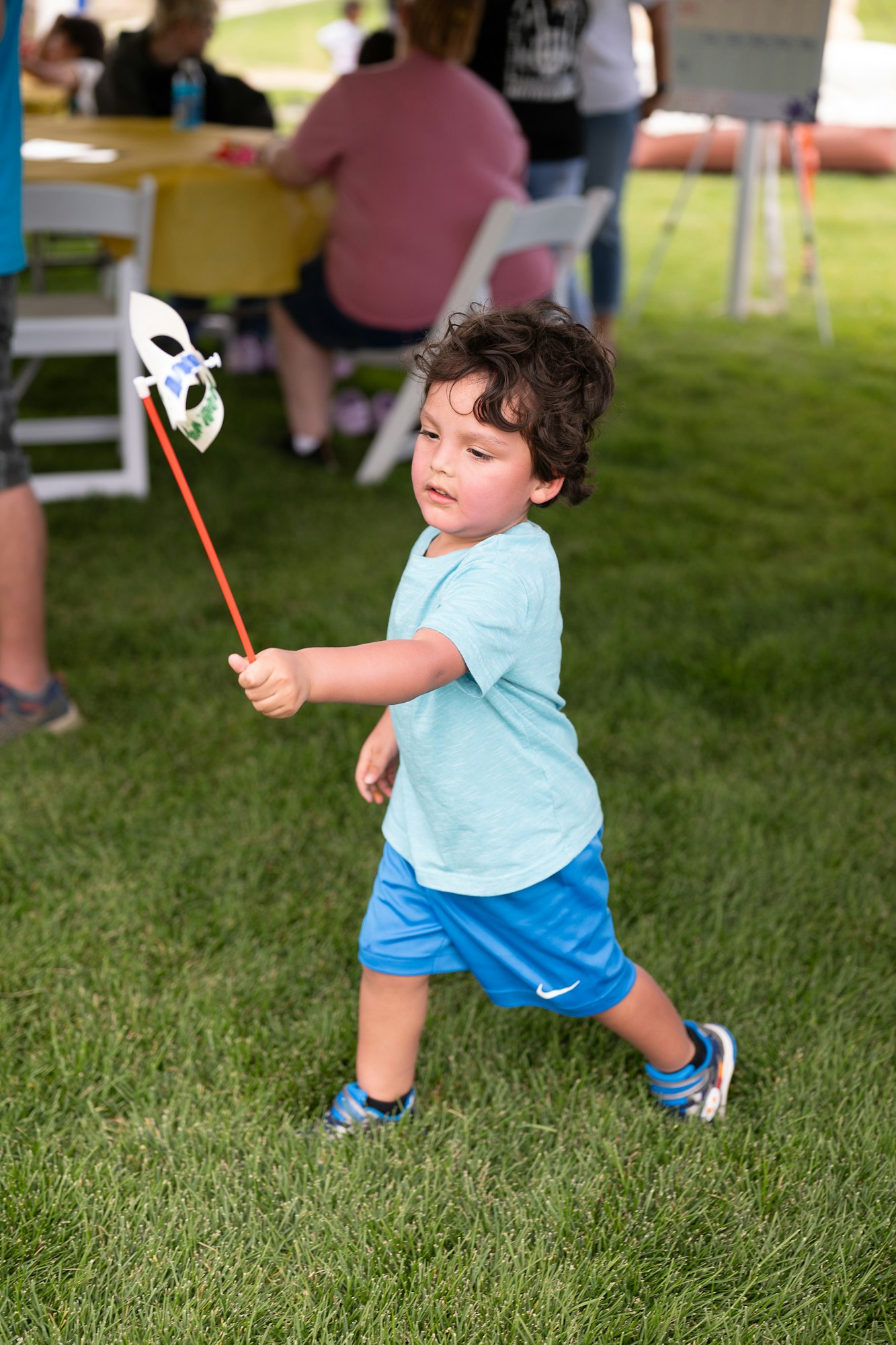Child walking on grass, holding a stick with a mask attached, at an outdoor event.