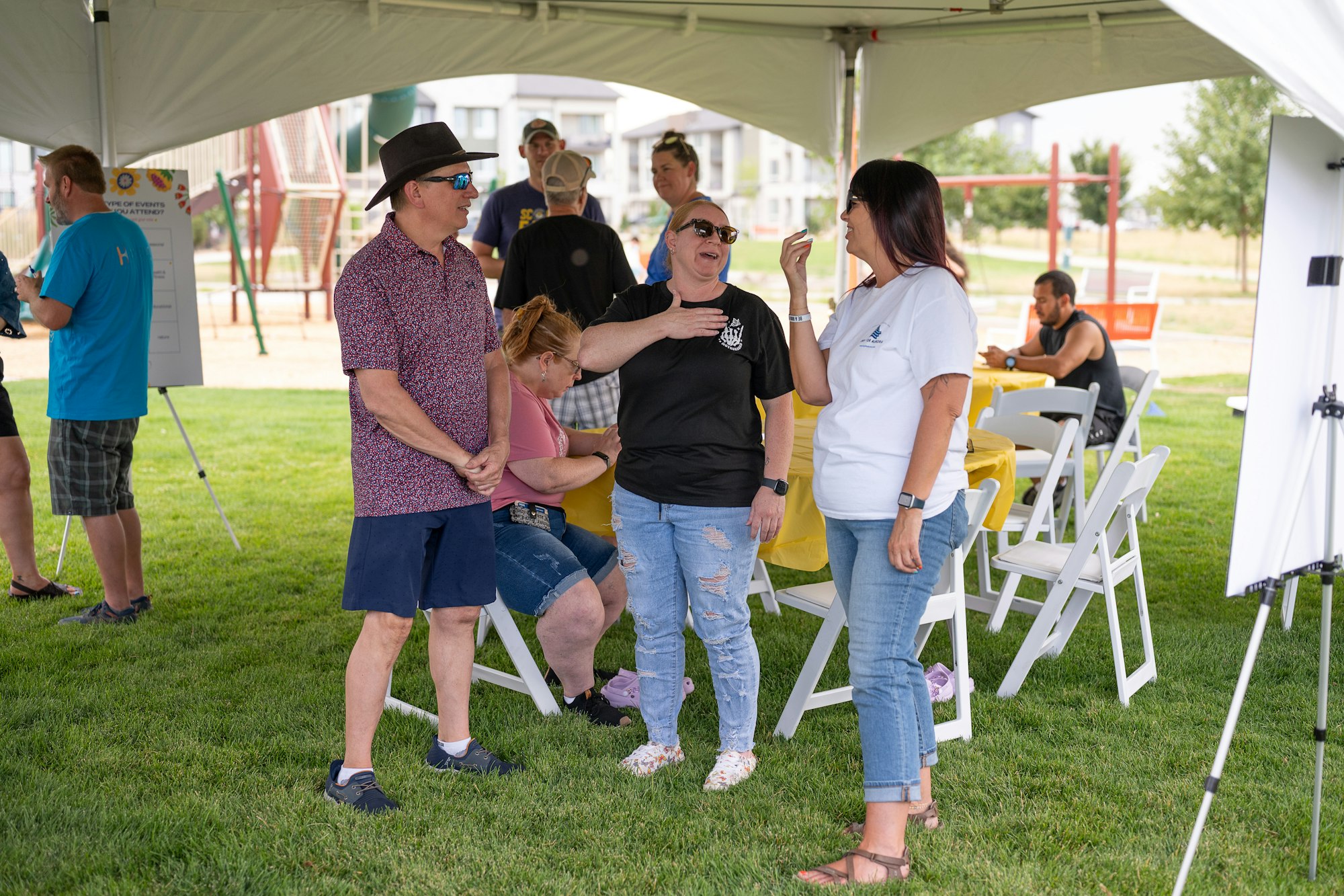 People chatting at an outdoor gathering under a tent with tables and chairs.