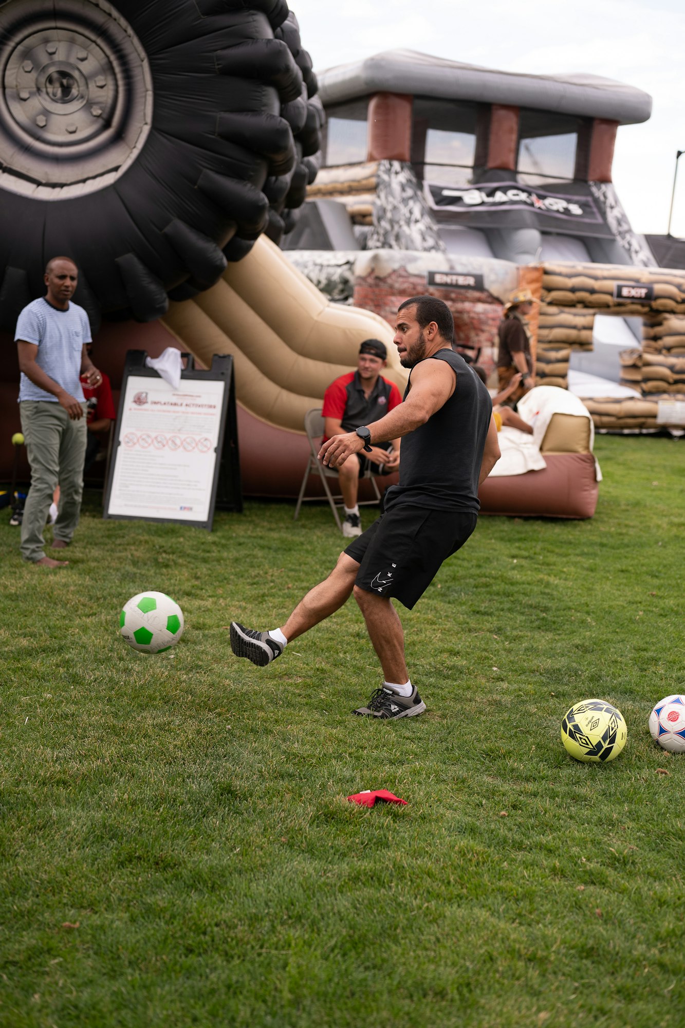 A man kicking a green soccer ball on grass with inflatable structures and people in the background.