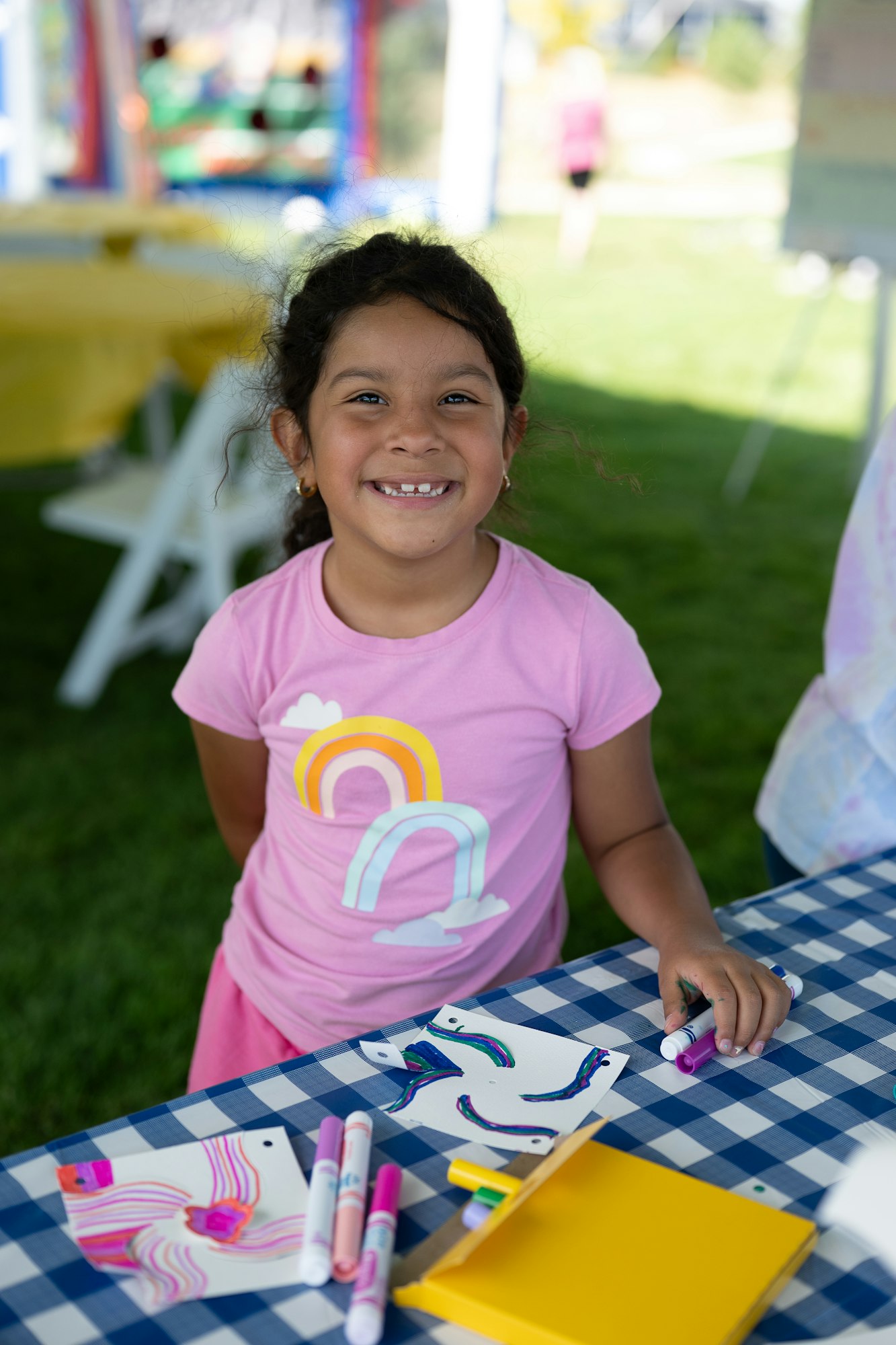 A smiling child in a pink shirt holds markers at a table with colorful drawings outdoors.