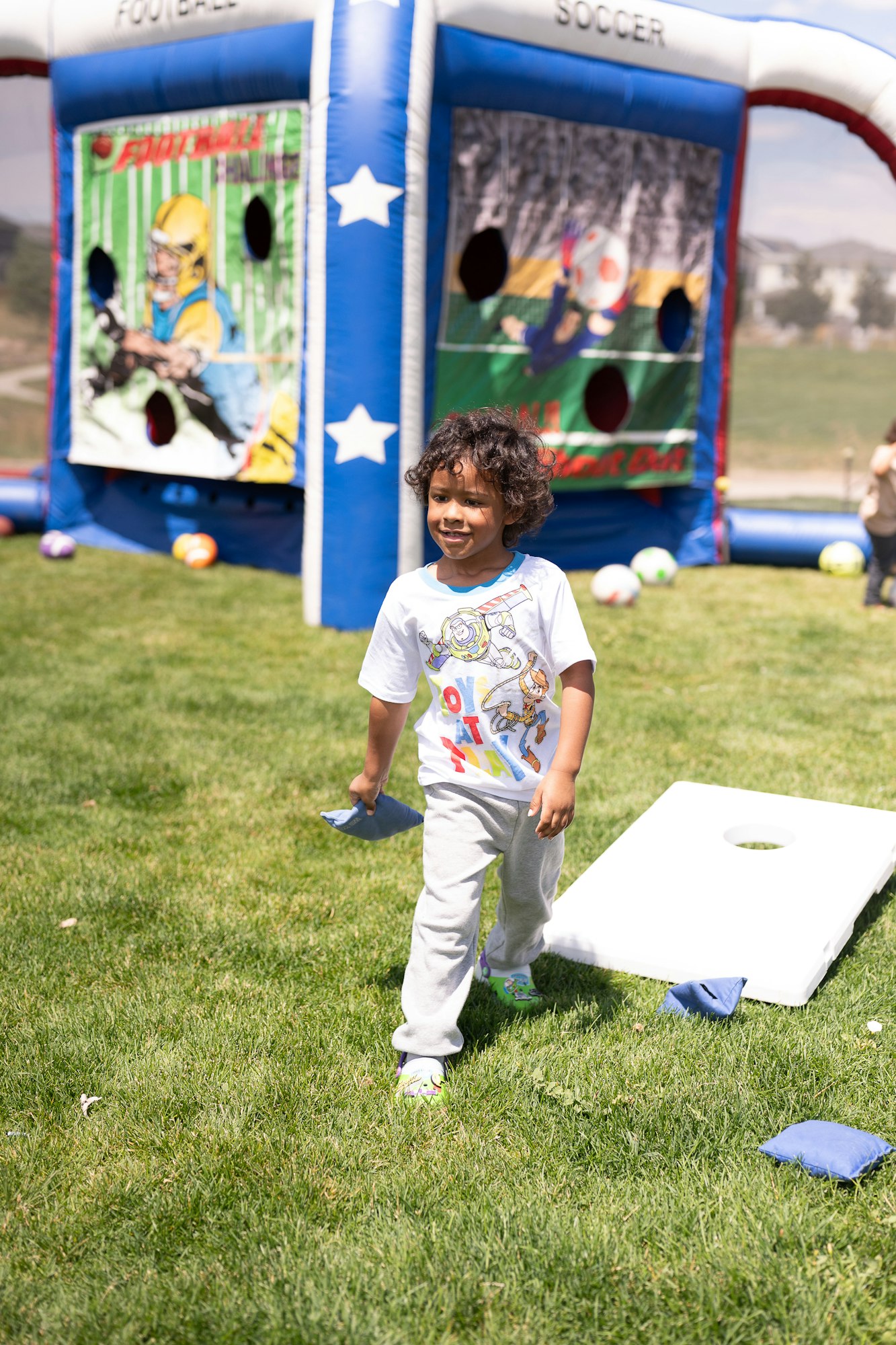 A child with a bean bag, a cornhole board, and an inflatable sports-themed backdrop outdoors.