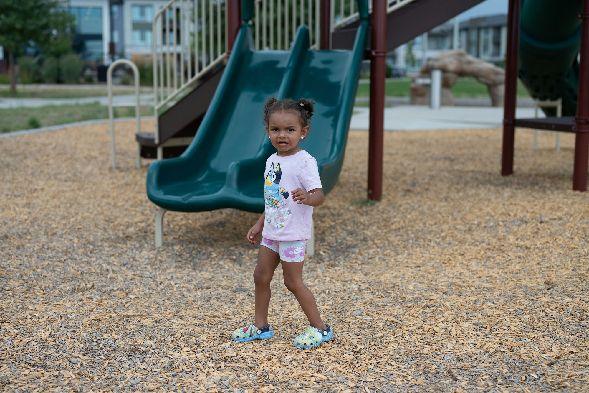 A child at a playground stands near a slide, wearing a T-shirt and shorts.