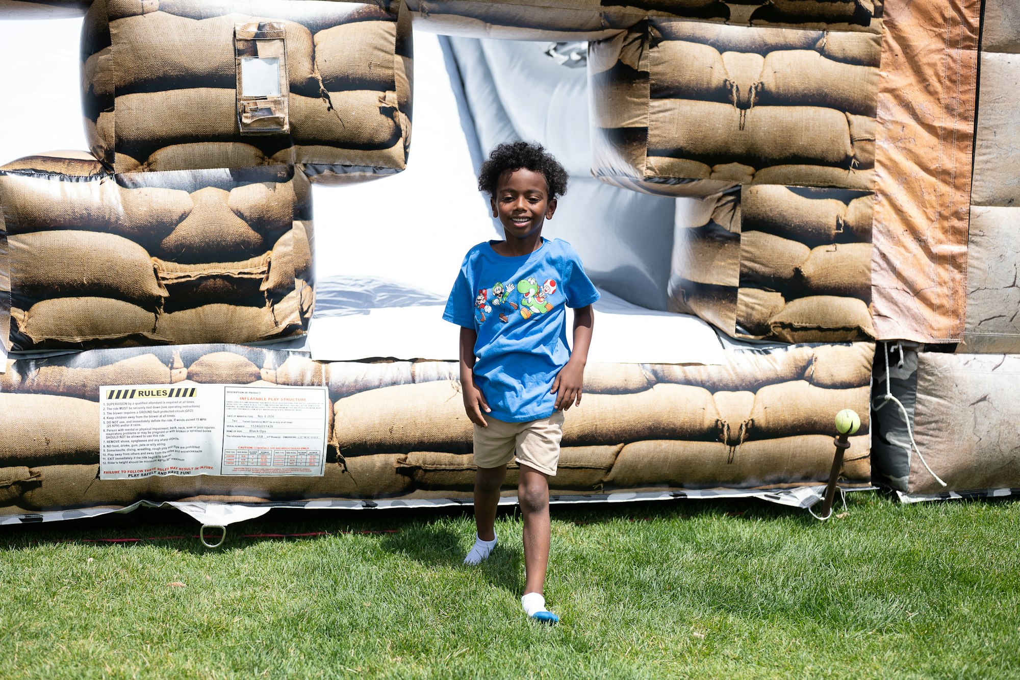 A child in a blue shirt walking outdoors in front of an inflatable bouncer with a sandbag pattern.
