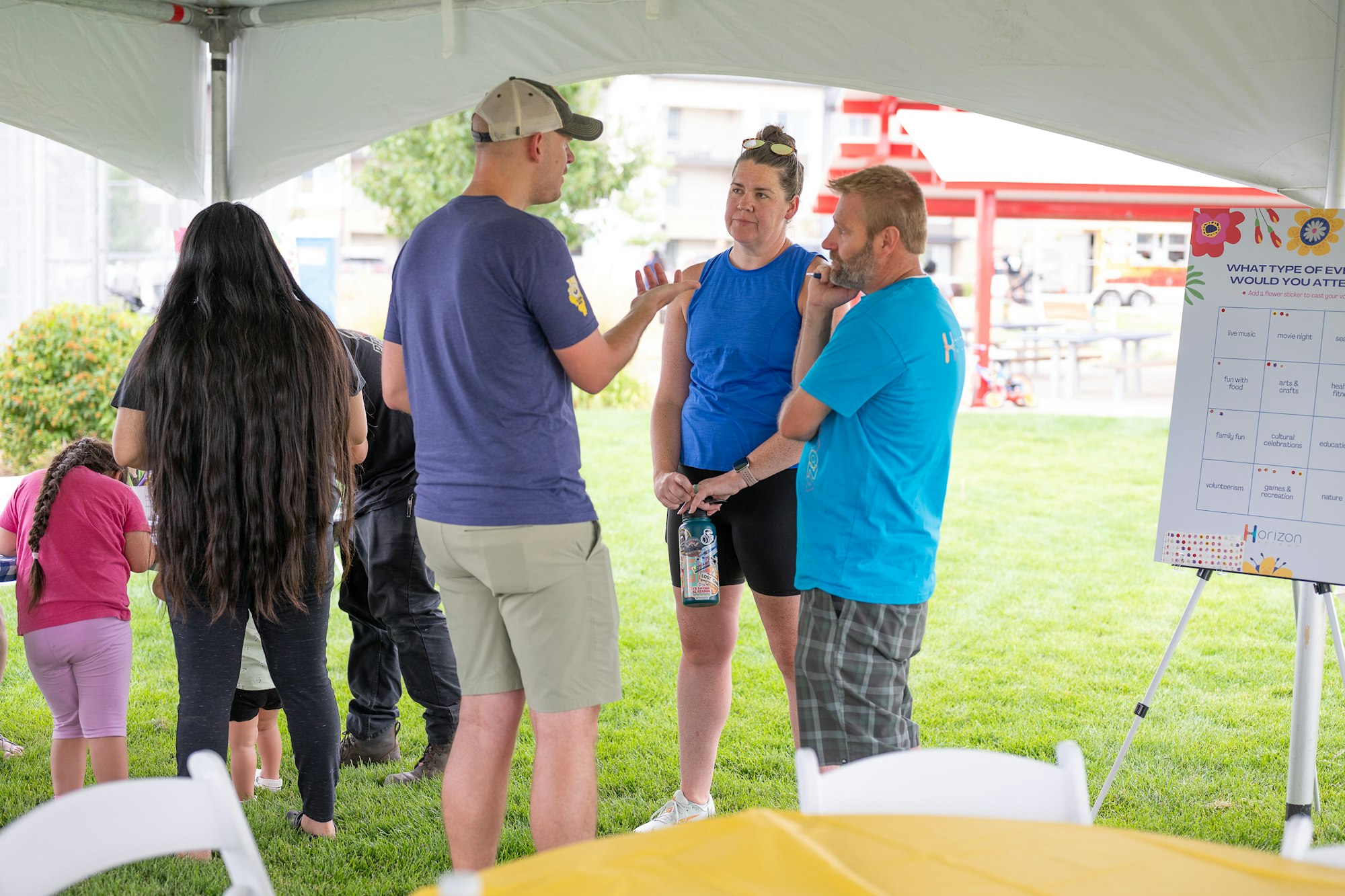 A group of people talking under a tent with a bingo game board in the background.