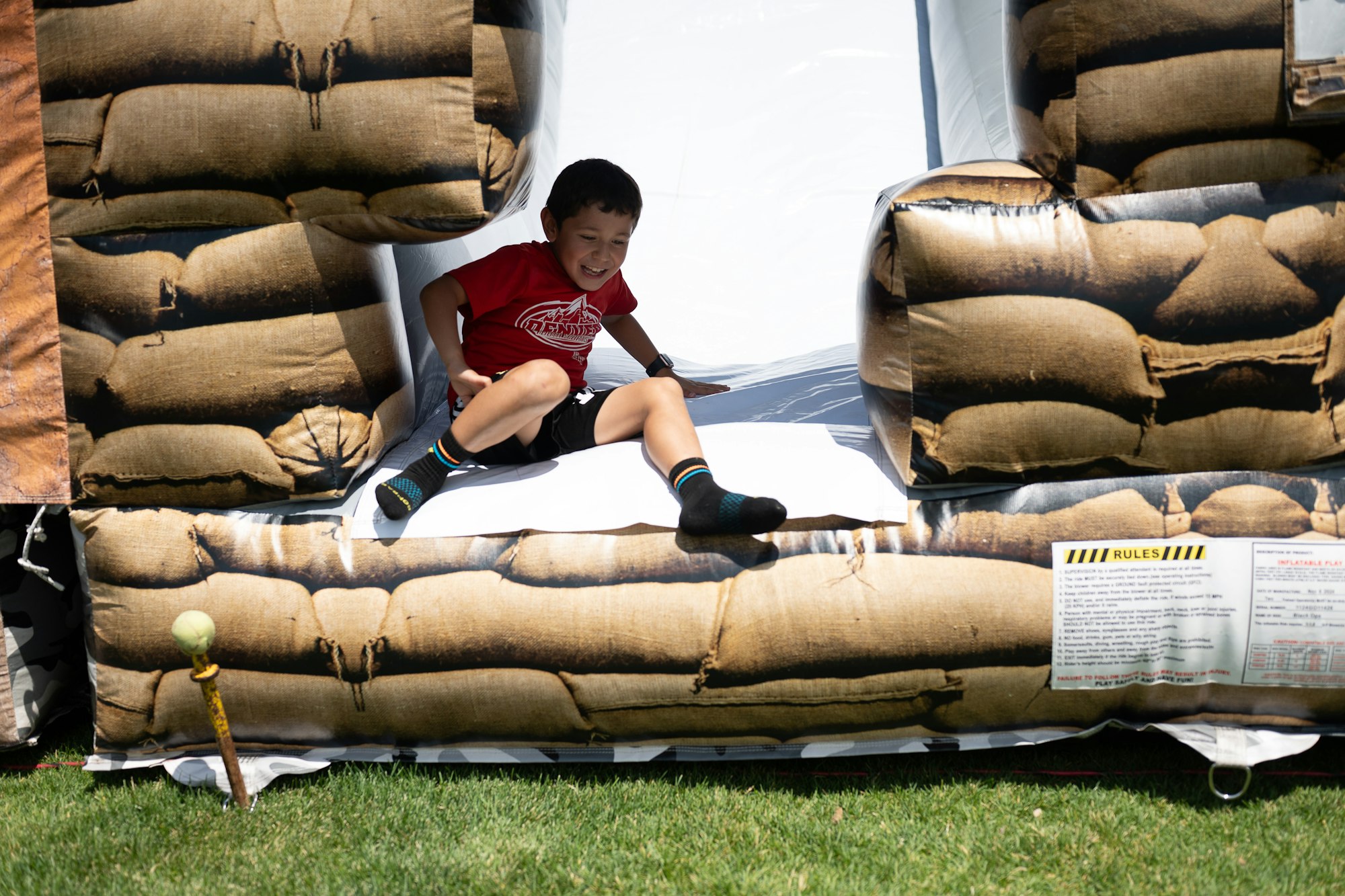 Child sliding out of an inflatable structure with a sandbag design on grass, smiling in a red shirt.