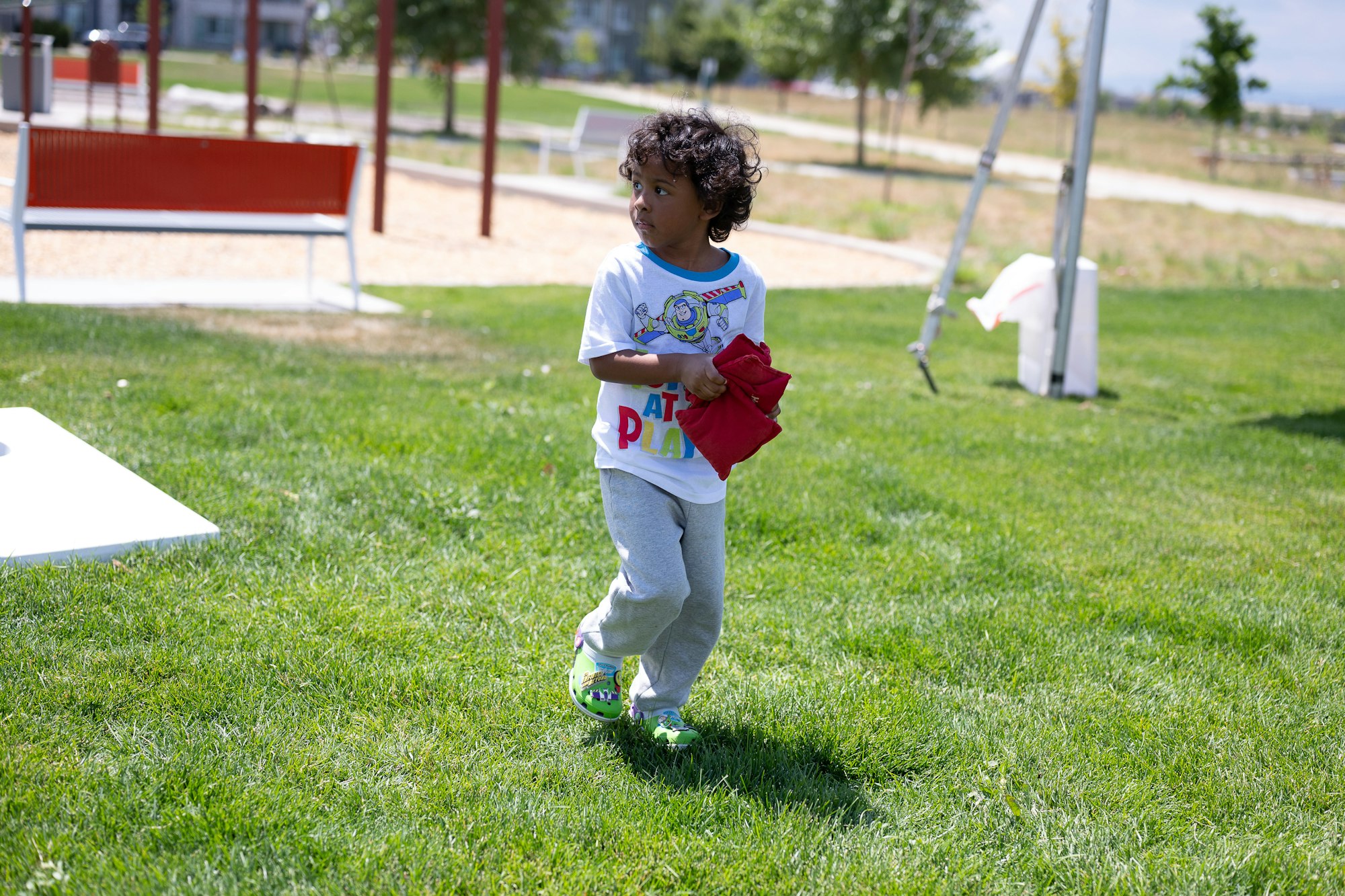 A child holding red bean bags in a park, wearing a cartoon-themed shirt and green shoes.