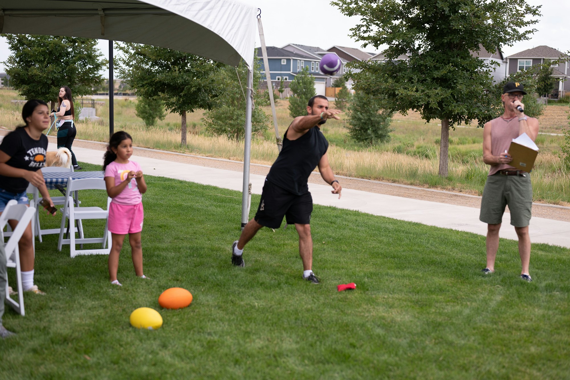 A group of people outdoors; one man throws a ball, others watch. A man with a mic and clipboard stands nearby.