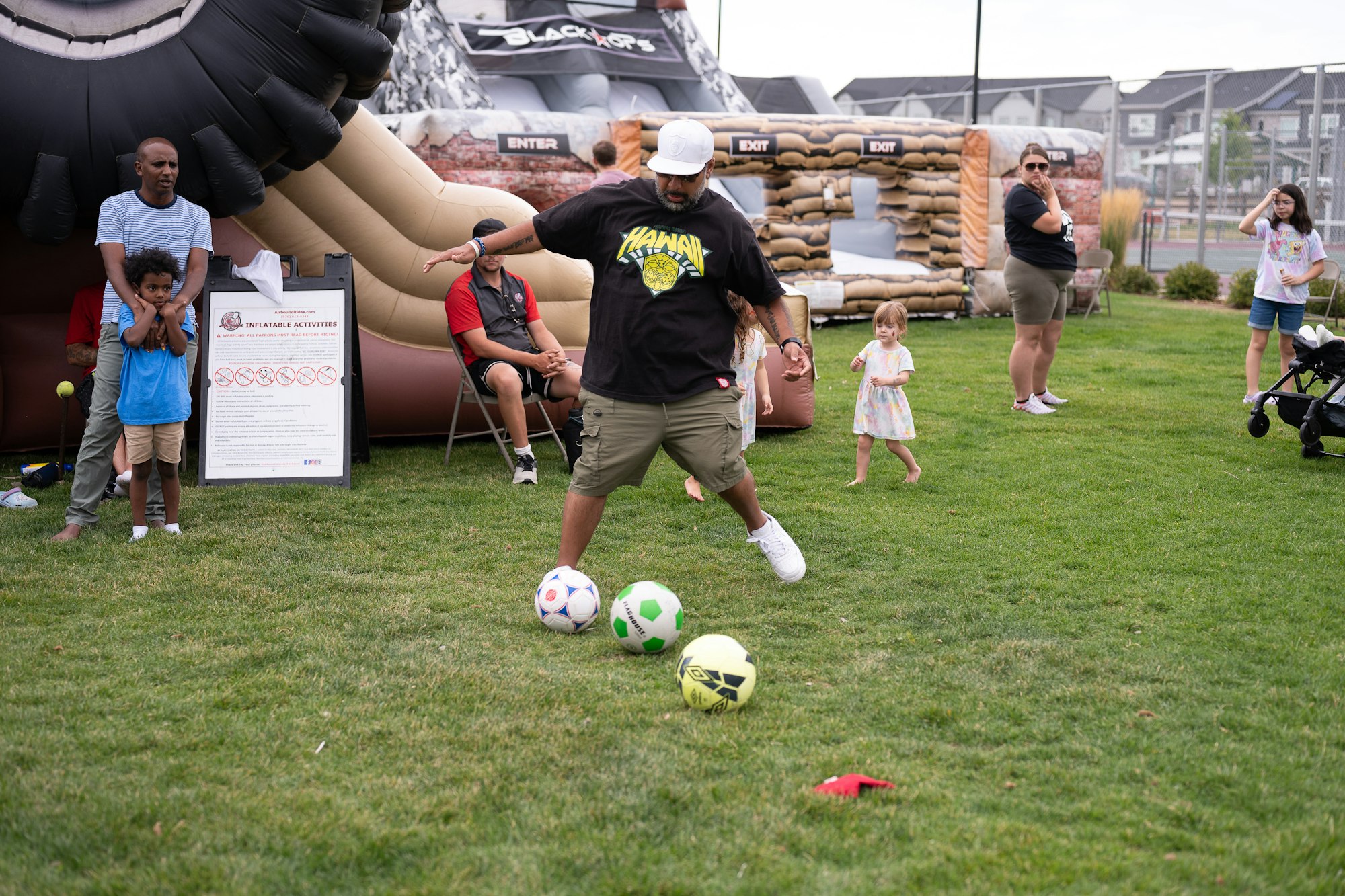 A man playing with soccer balls on grass at an outdoor event with inflatable structures and spectators.