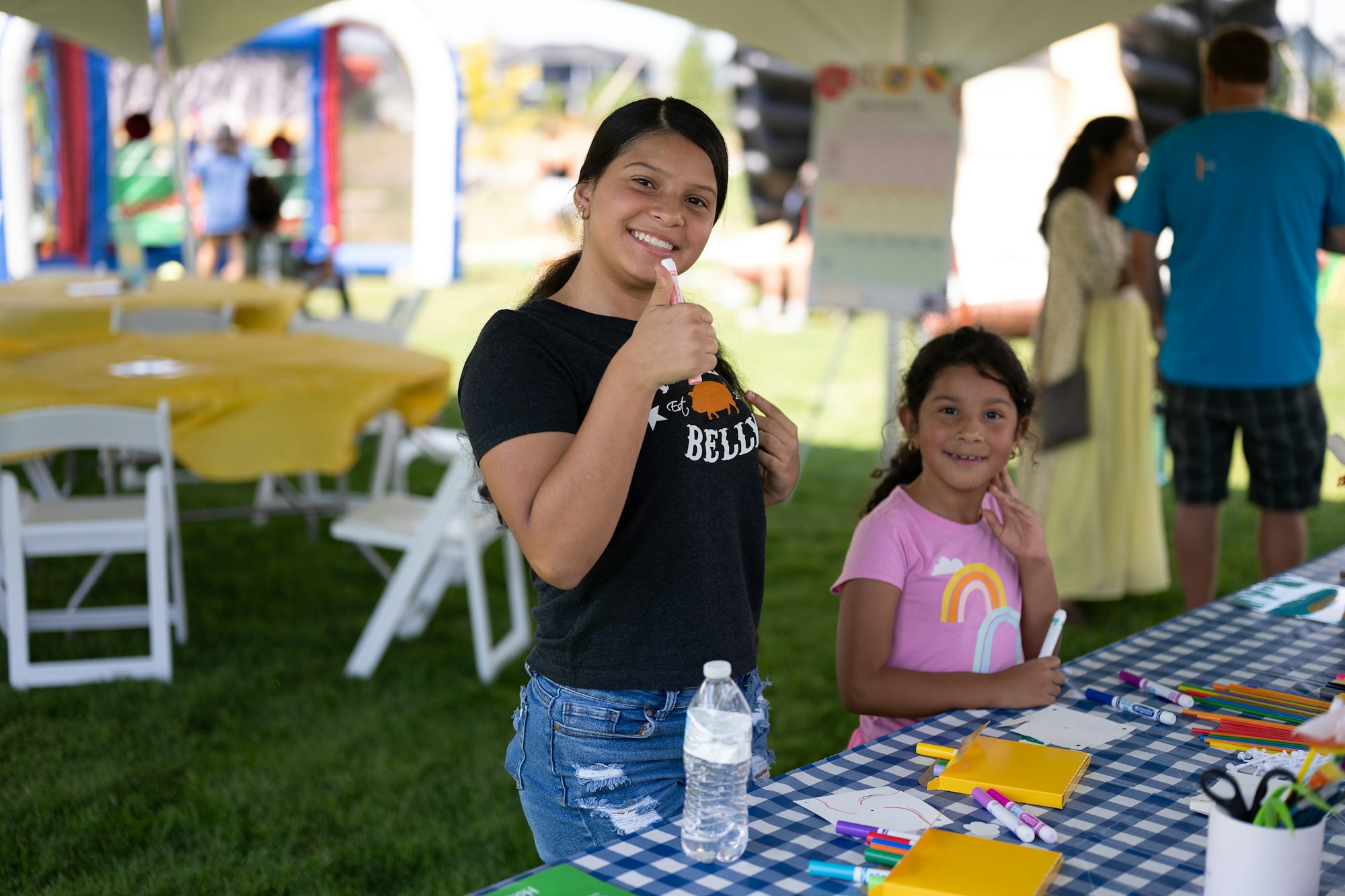 Two people smile at a table with art supplies under a tent at an outdoor event.