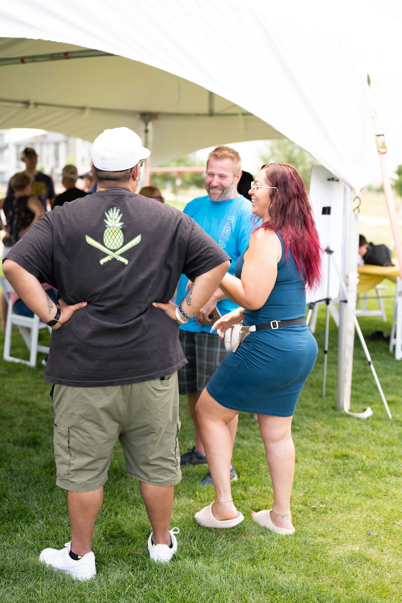 Three people are chatting and laughing outdoors under a tent. One wears a shirt with a pineapple design.