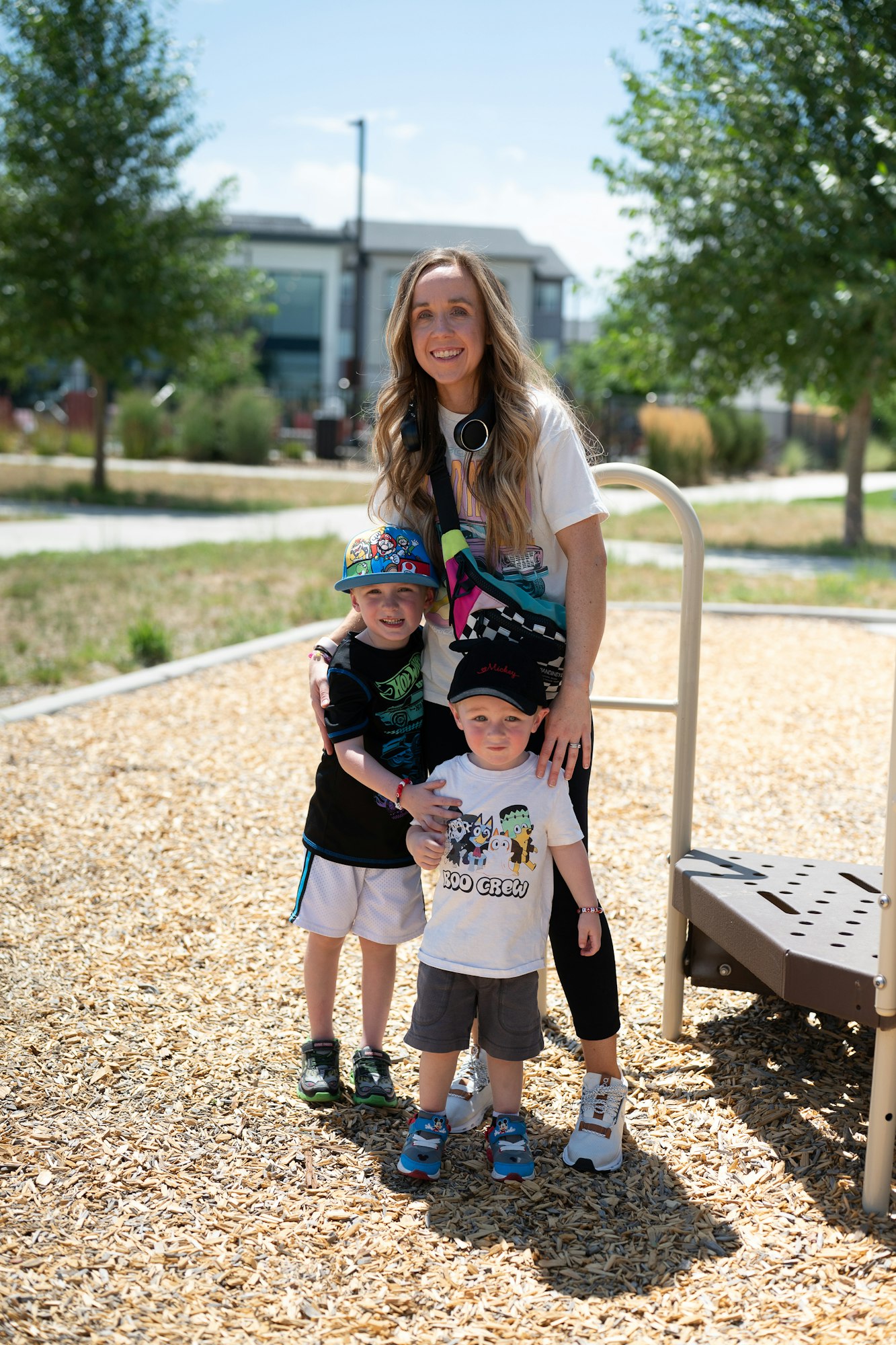 A woman and two kids smiling at a park, standing on wood chips with play equipment nearby.