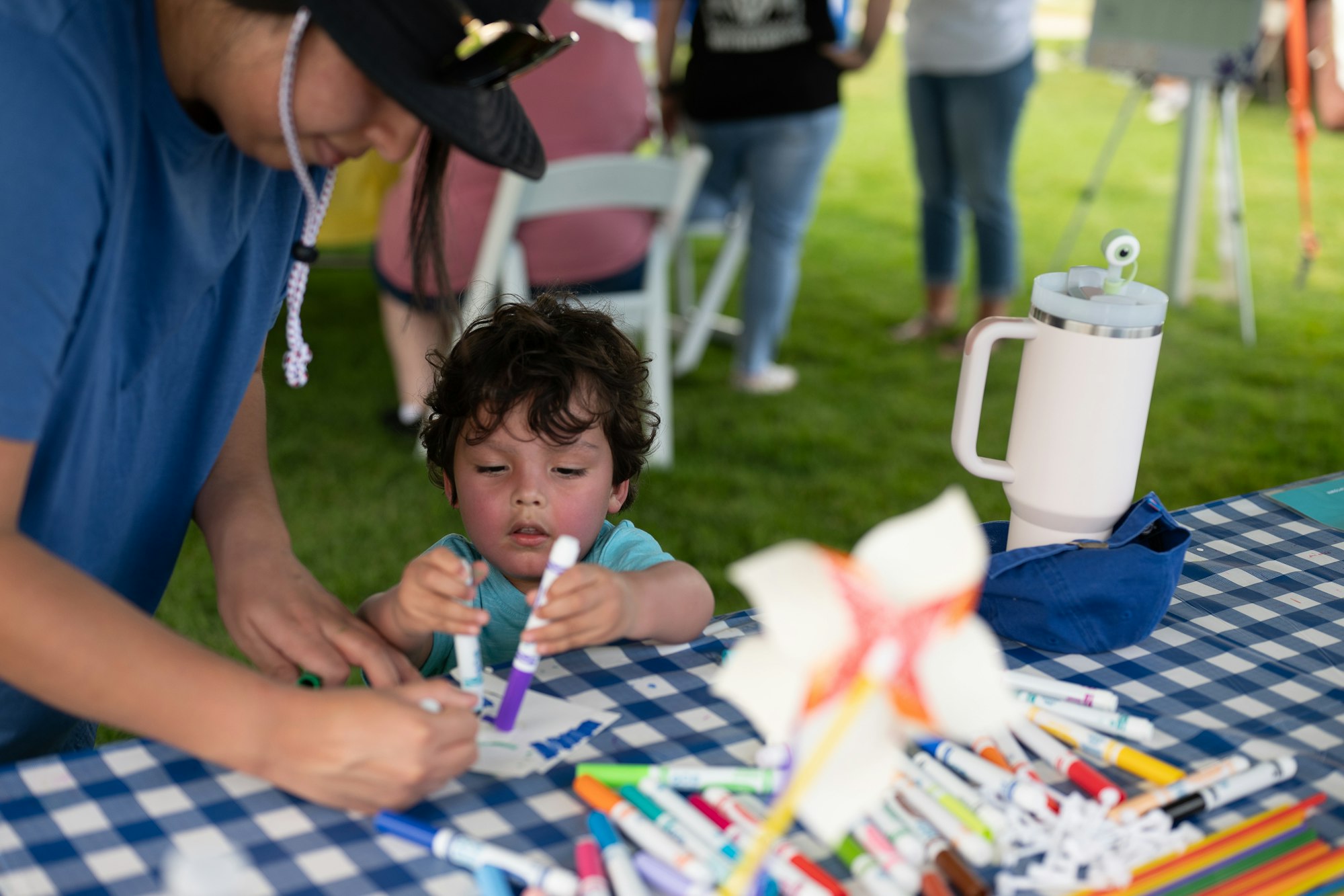 A child and an adult are engaged in a craft activity at a table with markers and a water bottle outdoors.